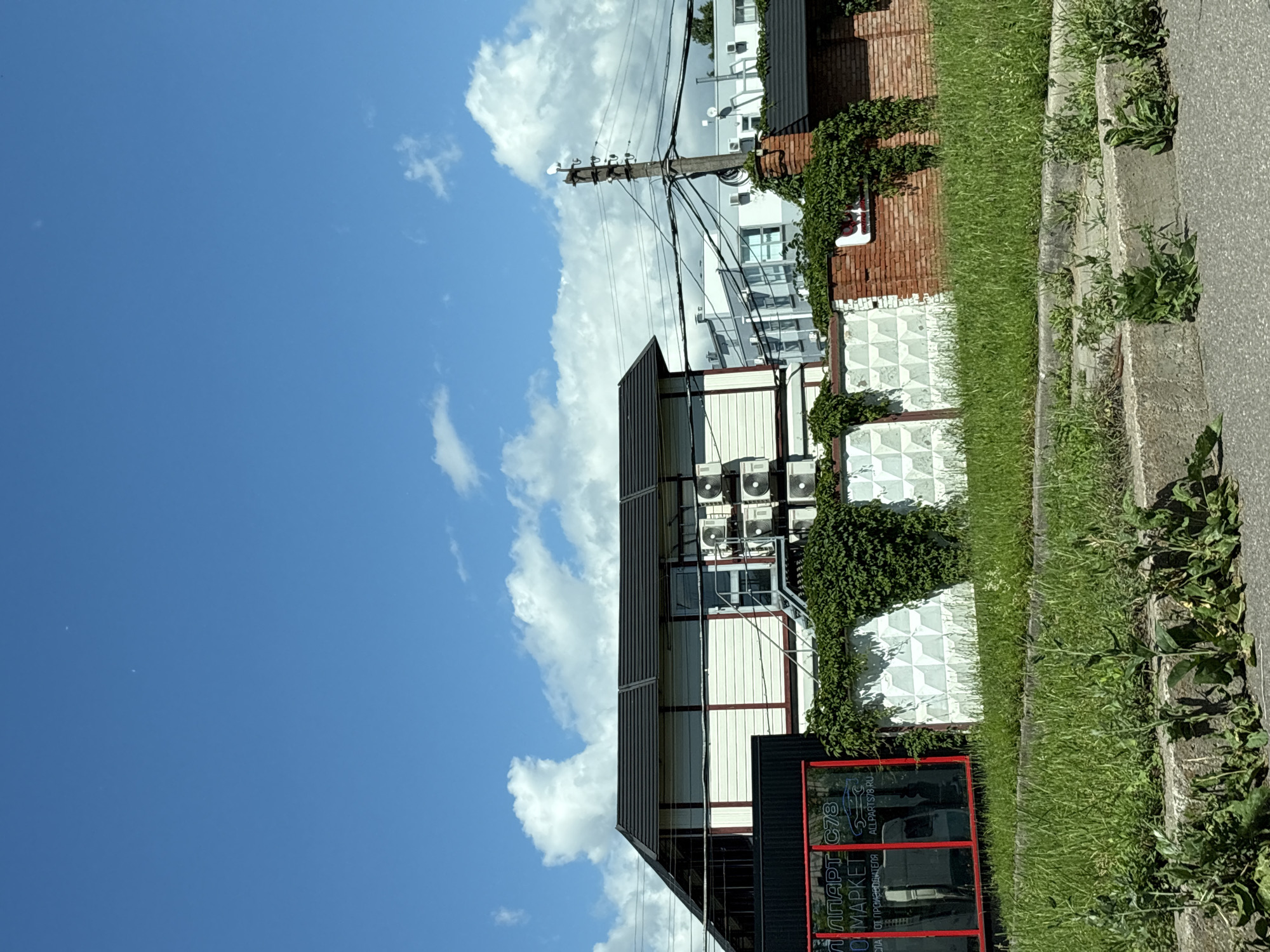 The image shows a bright, sunny day with a blue sky filled with fluffy white clouds. In the foreground, a patch of overgrown grass and weeds grows in front of a concrete curb and asphalt road. Behind the grass is a low, white wall with a repeating diamond pattern, partially covered in green ivy.
To the left of the wall, a building with a black roof and white siding trimmed in red is visible. A large red-framed window displays text in Cyrillic letters, reading "АППАРТ С78" and "О МАРКЕТ". The building has an exterior staircase leading up to it, and several air conditioning units are mounted on its side.
The wall continues to the right, transitioning into a red brick section that is also covered in ivy. A utility pole with multiple wires runs vertically through the middle of the scene, behind the building and wall. In the background, several other buildings, appearing to be commercial or industrial, are visible under the blue sky. The scene appears to be an outdoor commercial area.