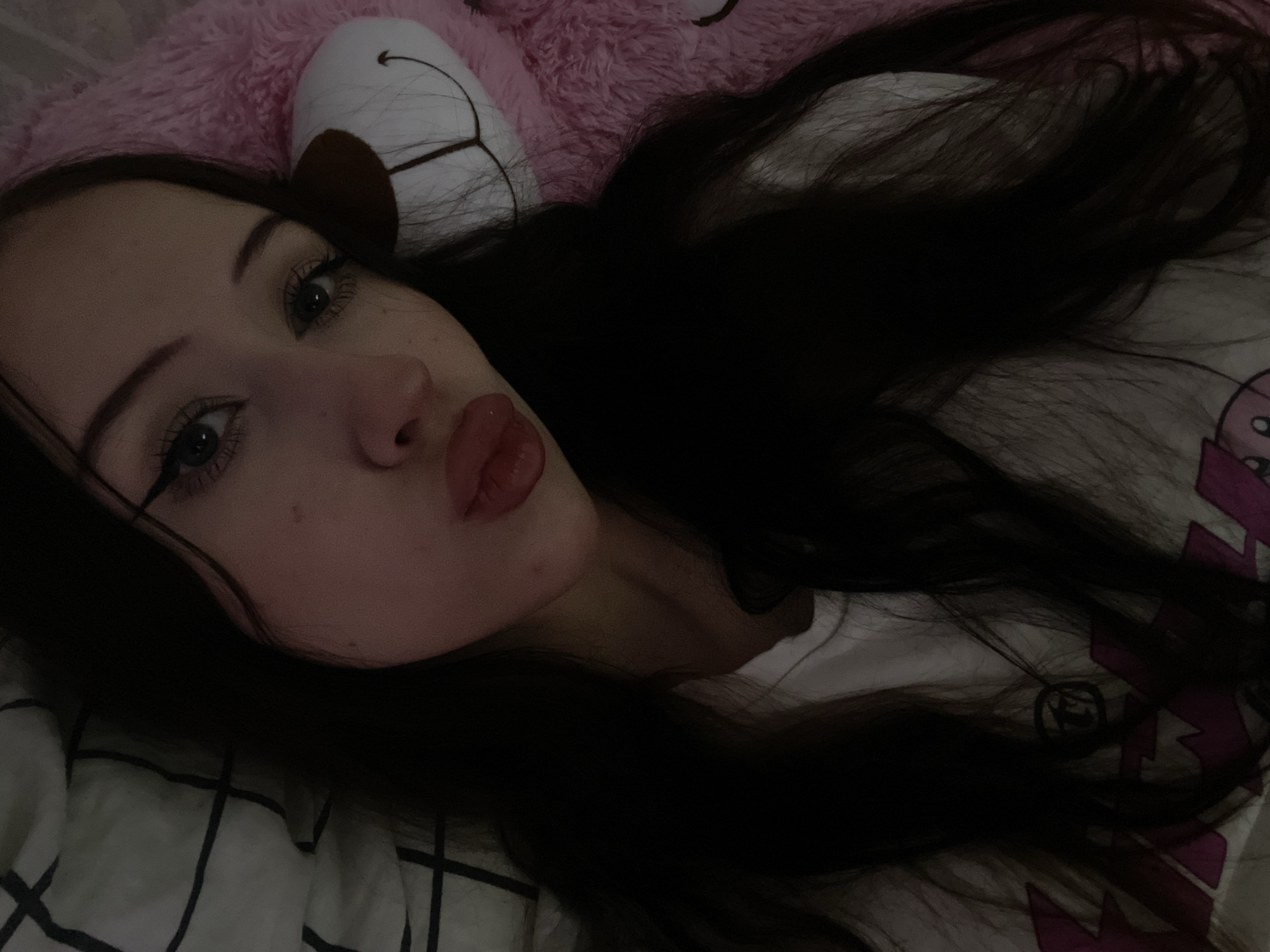 A young woman with long dark hair and dark eye makeup is lying down, her head resting on a black and white checkered pillow and a pink fuzzy object, possibly a stuffed animal. She is wearing a white t-shirt with a graphic design. Her lips are pursed. The lighting is dim, suggesting an indoor environment, possibly a bedroom. No specific details about the location in Votkinsk, Russia can be inferred from the image.