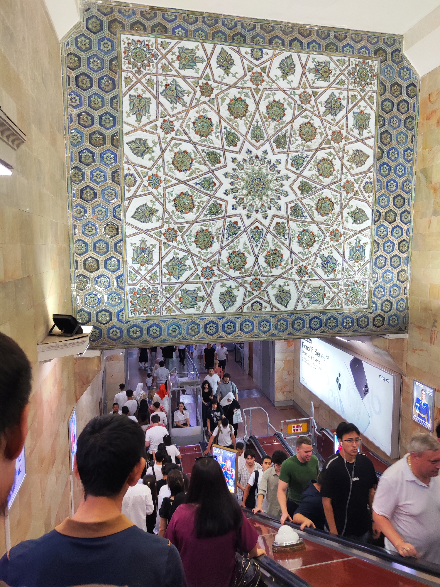 The image shows an interior view of a busy subway station escalator in Tashkent, Uzbekistan. The station is adorned with intricate blue and white geometric tilework on the walls and ceiling, featuring floral motifs and hexagonal patterns. A large, ornate tile mosaic is prominently displayed above the escalator, with a central starburst design surrounded by smaller floral elements.
A crowd of people, dressed in casual attire, are ascending the escalator. They appear to be locals and tourists, with a mix of ages and genders. Some individuals are looking at their phones, while others are engaged in conversation. An advertisement for Oppo Reno16 Series phones is visible on the wall beside the escalator, as well as another advertisement featuring a man in a blue and yellow sports uniform. The lighting is a mix of natural and artificial sources, illuminating the scene. The overall impression is one of a vibrant and bustling urban transportation hub.