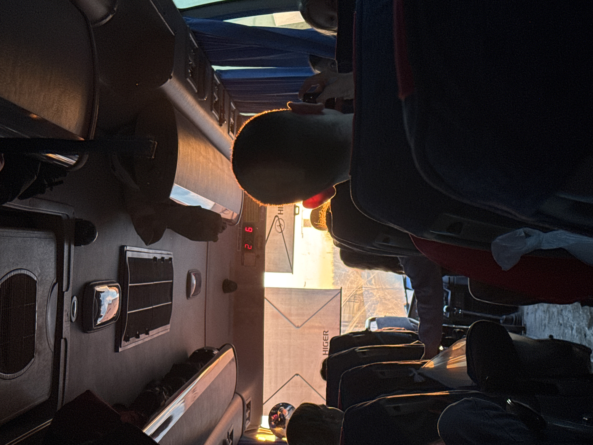The image depicts the interior of a Higer bus in Osa, Russia. Passengers are seated, with their luggage stored overhead. A digital clock displays "2:6." Light enters through the front window, illuminating the bus.