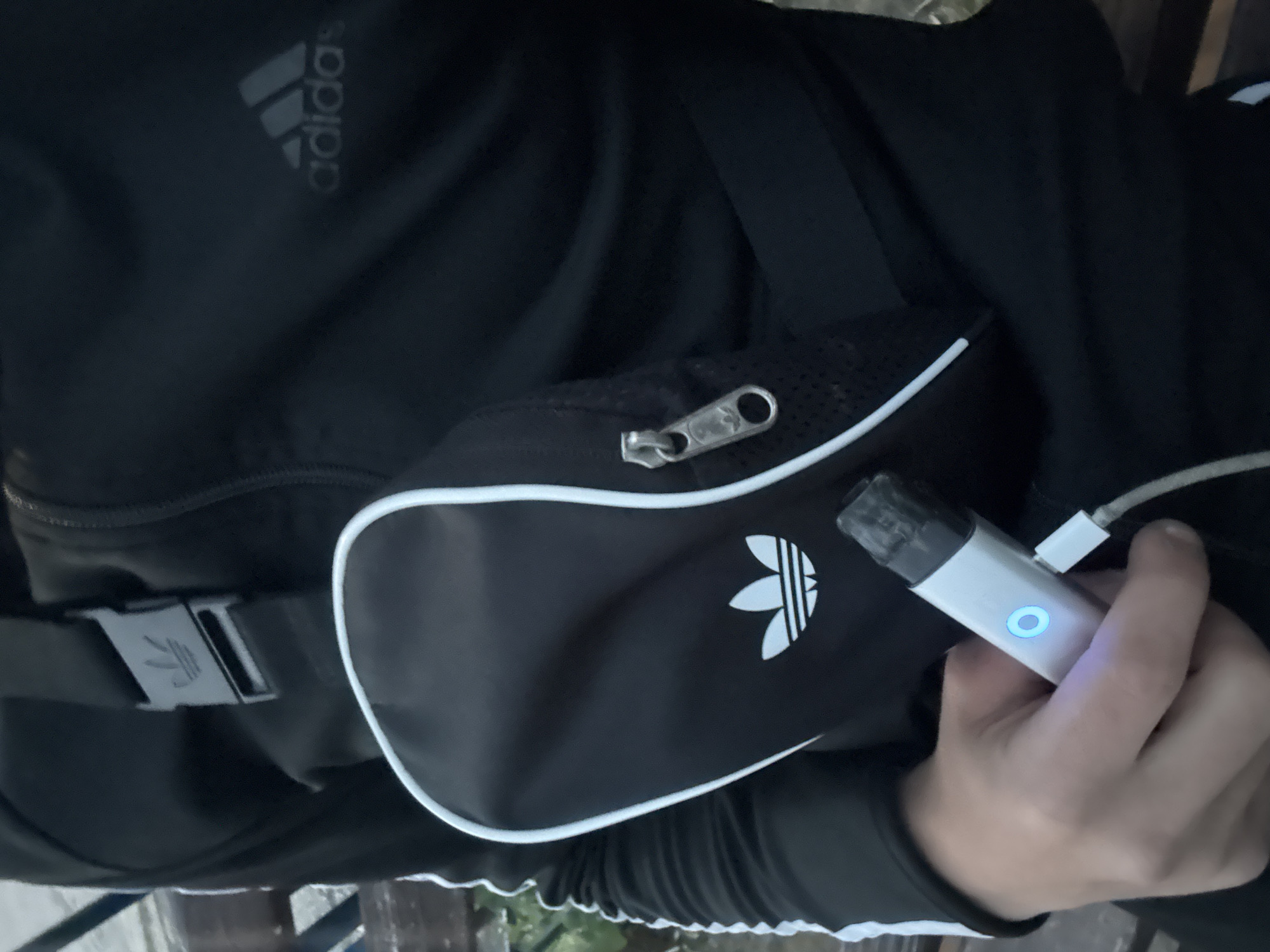 A person is wearing a black Adidas tracksuit and a black Adidas crossbody bag. The person is holding a vape pen connected to a charger in their right hand. The location is unknown, and there is no evidence to support it being in Varna, Bulgaria.
