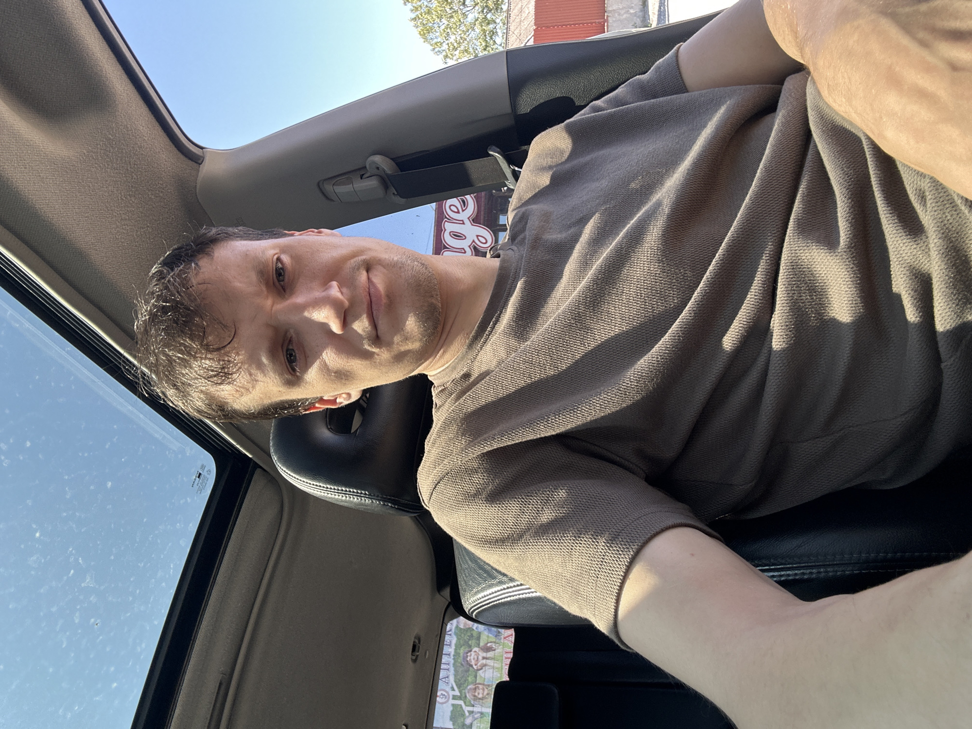 A man is sitting in the driver's seat of a car, looking at the camera. The car's sunroof is open, and the sky is visible. The man is wearing a brown t-shirt. In the background, a building with a red roof and the word "age" in large letters is visible. There is also a tree and a red fence. A sticker with the image of several children is visible on the rear window of the car. The image was taken in Shatava, Ukraine.