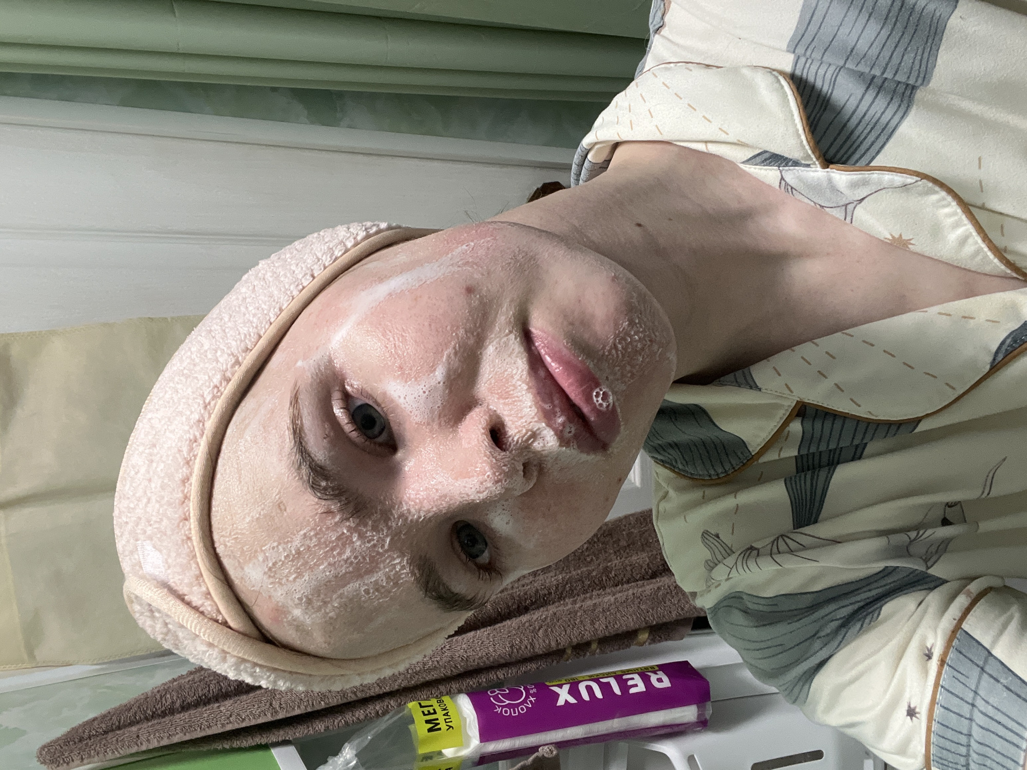 A close-up, slightly tilted indoor portrait captures a person engaged in a skincare routine. The subject, who appears to be a young individual with fair skin and light-colored eyes (blue/grey), looks directly at the viewer with a neutral expression. Their face is generously covered in a white, foamy product, consistent with a cleanser or face mask, and a small white bubble rests on their upper lip.
They are wearing a soft, light pink or peach-colored terry cloth headband, which keeps their hair away from their face. Their attire is a set of patterned pajamas, featuring a cream or off-white base with distinct grey-blue stripes or abstract shapes, accented with occasional orange-brown piping or details.
In the foreground, a folded brown towel is visible beneath the person's head. On a white surface to the right of the towel, likely a shelf or stool, sits a purple and white package. The brand name "RELUX" is clearly visible in white letters, and below it, Cyrillic text reads "ХЛОПОК" (KHLOPK, meaning cotton) and "100%", indicating a cotton product. Adjacent to it, a partially obscured and blurry package with yellow and green accents shows fragments of Cyrillic text, including "МЕТ" and "УПАКОВ", likely part of a product description.
The background consists of light-colored, possibly off-white, vertical paneling, suggesting an indoor setting such as a bathroom or bedroom. A horizontal green or teal element is visible at the very top of the frame. The overall impression is one of a personal moment of self-care. Given the visible Cyrillic text, the location context provided, Chaikovsky, Russia, is consistent with the visual cues.
