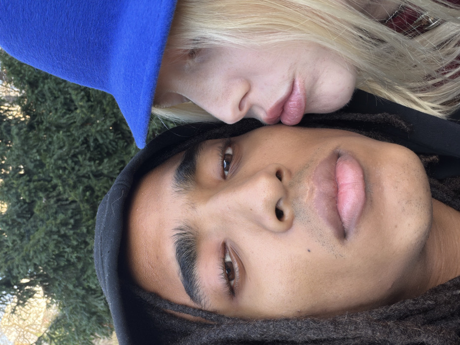 The image displays a close-up portrait of two individuals in an outdoor setting. The background is composed of blurred green foliage, consistent with trees or dense bushes, under natural daylight.
The individual on the left has dark brown skin, dark dreadlocks, and wears a black hooded garment. Their brown eyes are open and directed towards the camera. Features include a broad nose, full lips, and minimal visible facial hair on the upper lip and chin.
The individual on the right has light skin and blonde hair, partially obscured by a bright blue, wide-brimmed felt hat. This individual's face is presented in profile, oriented towards the left subject. Their lips are visibly pursed into a "kissing" configuration, positioned in close proximity to the left subject's right cheek area. A fragment of a red garment or accessory is visible near the lower right edge of the frame.
The precise location within Paris, France, is not discernible from the visual evidence provided.
