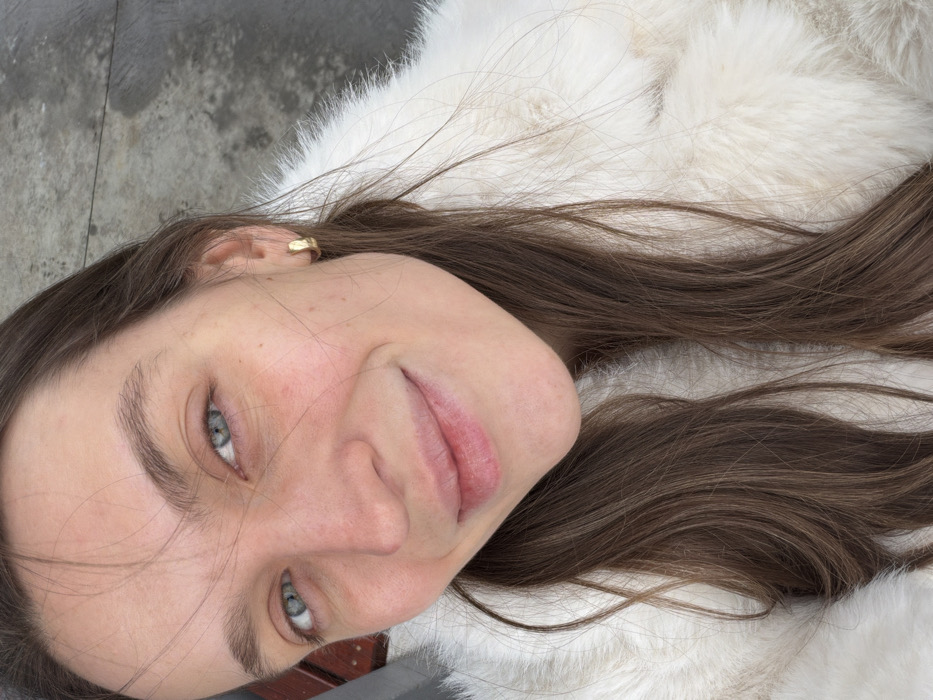 A woman with fair skin and brown hair lies against a white, furry surface. Her face is angled towards the camera, showcasing her light blue eyes and pink lips. She wears a small gold hoop earring. The background is a mix of concrete and a glimpse of wood, suggesting an outdoor setting. Her expression is subtle, hinting at a relaxed or contemplative mood.