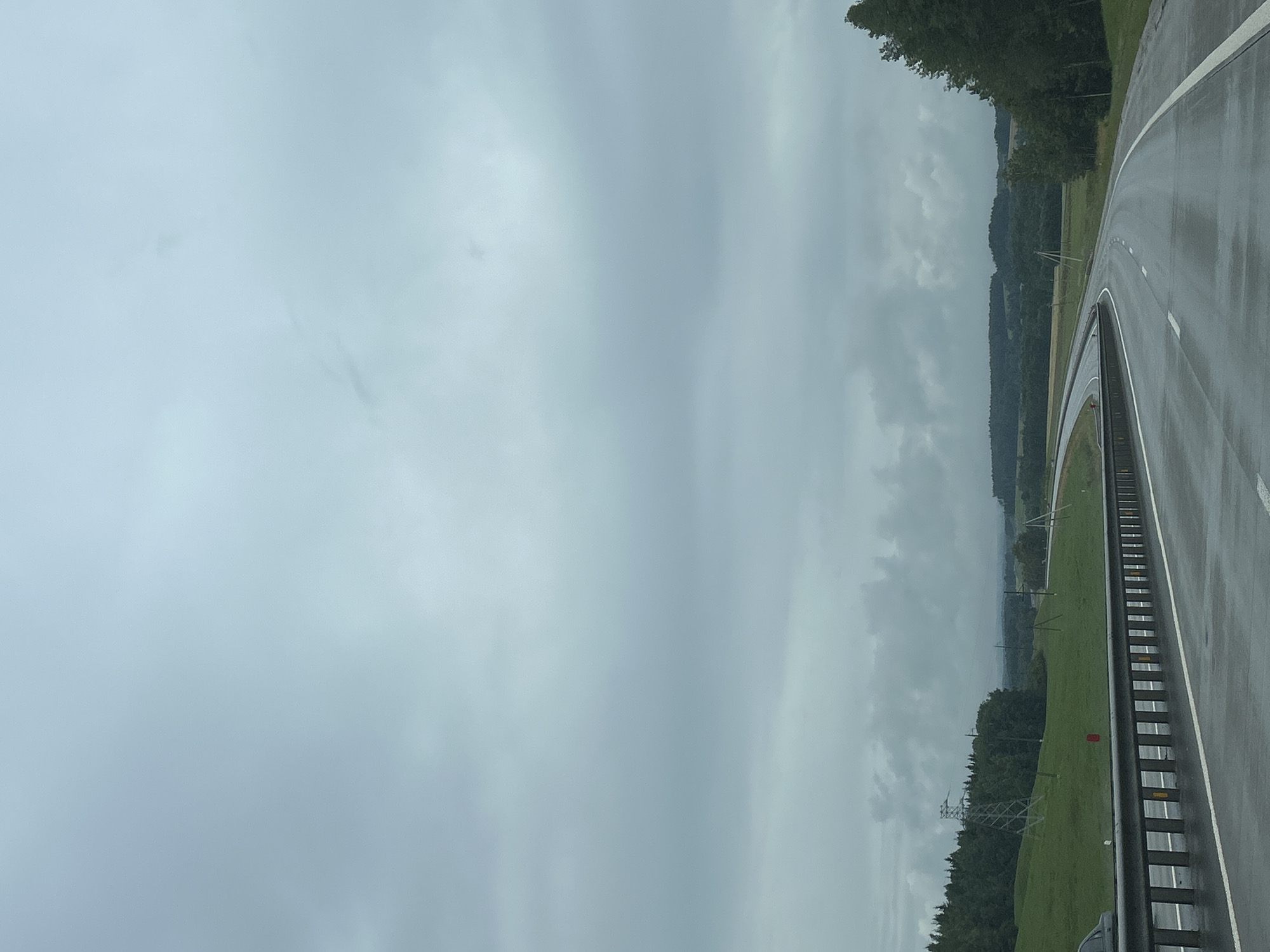 This image depicts a wet, curved highway in a rural, possibly forested area. The sky is overcast with heavy clouds. A guardrail runs along the left side of the road, with a white line separating the lane. In the distance, there are rolling green hills and a dense line of trees. A transmission tower is visible on a grassy slope to the left. The road surface appears reflective, indicating recent rain. The location is described as Lahojsk Selsaviet, Belarus. There are no people visible in the image.