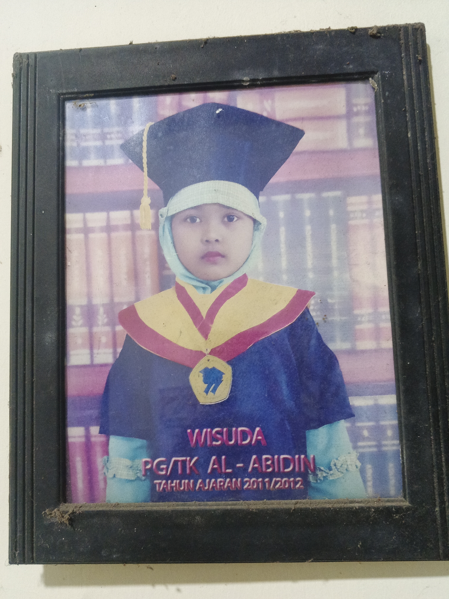 A framed portrait shows a young child wearing a graduation cap and gown. The child is wearing a light blue headscarf under the graduation cap, and a gold and red stole over the blue gown. A medal is hanging from the stole. The background of the portrait appears to be a bookshelf. Text on the portrait reads "WISUDA PG/TK AL-ABIDIN TAHUN AJARAN 2011/2012". The frame is black and appears to be dusty. The image was taken indoors, likely in a home or school environment in Penambangan, Indonesia.
