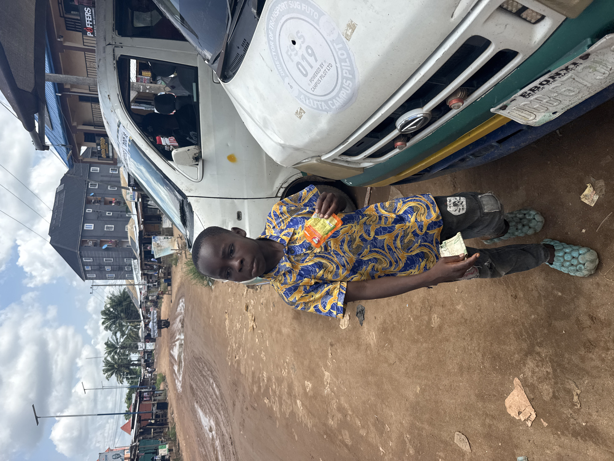 The image captures a young boy standing beside a white commercial minivan or bus on a dusty, unpaved road in Ihiagwa, Nigeria, under a bright, partly cloudy sky. The scene depicts a typical street environment in a semi-urban or rural area.
The boy, the central subject, wears a vibrant blue and yellow patterned short-sleeved shirt, dark pants with visible wear, and light blue, textured bubble-patterned slip-on shoes. He holds what appears to be a small, orange-and-yellow packaged snack in his left hand and several folded Nigerian Naira banknotes in his right hand. His expression is neutral to slightly serious as he looks towards the camera.
The white minivan, parked next to the boy, features several stickers and markings. On its front hood, a prominent circular sticker reads "DIRECTOR OF TRANSPORT SUG FUTO," with "019" in the center, and "POWERED BY CAMPUS PILOT LTD" and "ALUTA CAMPUS PILOT" also visible. The vehicle has a green and yellow stripe below the grill, and its license plate is partially visible, showing "EBONYI," "GOD IS THE NATION," "FEDERAL REPUBLIC OF NIGERIA," and numbers including "088-673."
In the background, two-story buildings line the street, with a shop sign "POFFERS" visible on one. Several palm trees and utility poles indicate a local streetscape. The ground is a reddish-brown dirt road, with some debris scattered about. The activity appears to be an everyday interaction, possibly a transaction like buying a snack or waiting for transport. The bright daylight suggests it is midday or early afternoon.