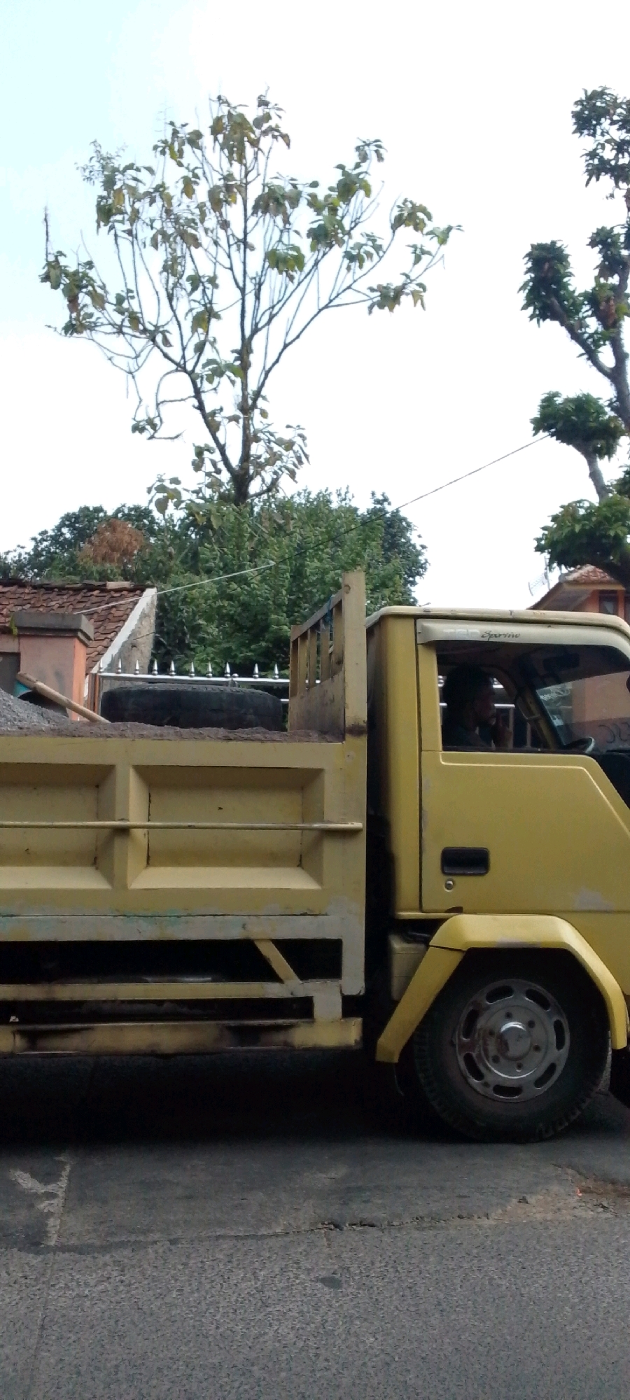 A yellow dump truck is parked on a paved road in the city of Cilaku, Indonesia. The truck is loaded with grey gravel in its bed. The driver, a man with dark hair, is visible in the cab, looking forward and holding the steering wheel. The truck has a visible front wheel with a silver rim. Behind the truck, a fence with pointed metal spikes separates the road from a property with a tiled roof house and dense green trees. The sky above is clear and bright.