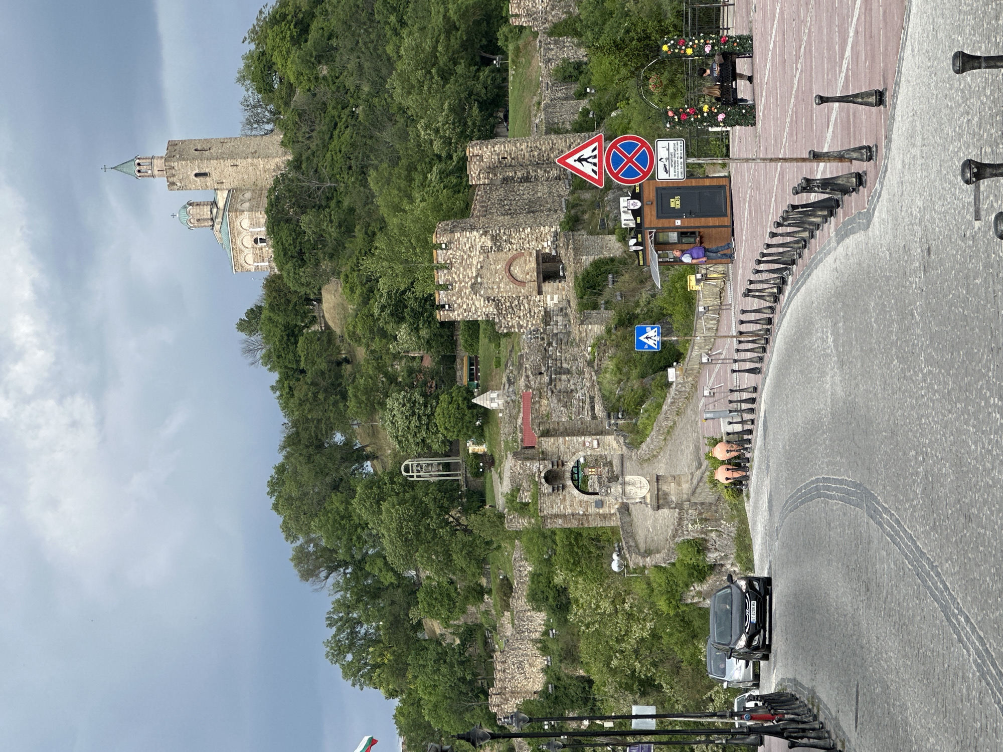 This image shows a view in Veliko Tarnovo, Bulgaria. A historic stone tower with a church sits atop a hill covered in dense green vegetation. Below, a section of the ancient fortress is visible with stone walls and archways. A paved walkway leads up the hill, marked with pedestrian crossing signs and other road signage. A ticket booth is located near the walkway, and a person is lying on a bench nearby. A paved road with decorative bollards runs alongside the walkway. Several cars are parked on the side of the road. The sky is overcast.