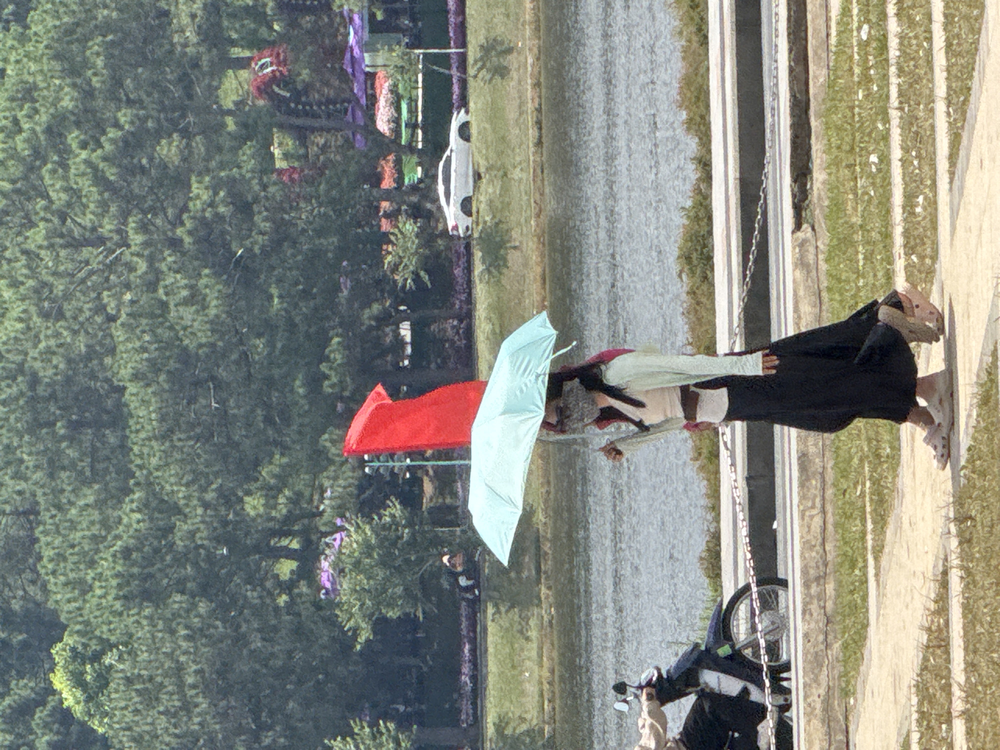 The image depicts a daytime scene in Da Lat, Vietnam, near Xuan Huong Lake. A woman walks on a paved pathway, holding a light blue umbrella over her head. She is wearing a light-colored top, a long black skirt, and a face mask. Her right hand is holding the umbrella handle, and her left hand is near her side. Behind her, a red flag is visible, fluttering in the breeze, and a motorcycle is partially visible on the left side of the frame. In the background, Xuan Huong Lake is present, with a white car driving on a road along the embankment. The environment includes trees and lush greenery, suggesting a park or scenic area. The overall impression is a casual outing in a public space.