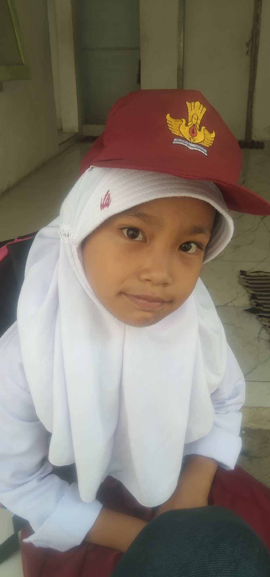 A young Indonesian girl, appearing to be of primary school age, is captured in a close-up, waist-up portrait. She is wearing a white hijab and a white school uniform shirt, partially obscured by the hijab. A maroon cap with a gold emblem featuring a bird of paradise is perched on her head, covering the top of her hijab. Her gaze is directed slightly to the left of the camera, and she has a neutral to slightly smiling expression. The background suggests an outdoor or semi-outdoor setting, possibly a schoolyard or porch area, with a light-colored wall and floor. A small, dark rug is visible on the floor to her right. The lighting is even, indicating daylight. The overall impression is that of a candid snapshot.
