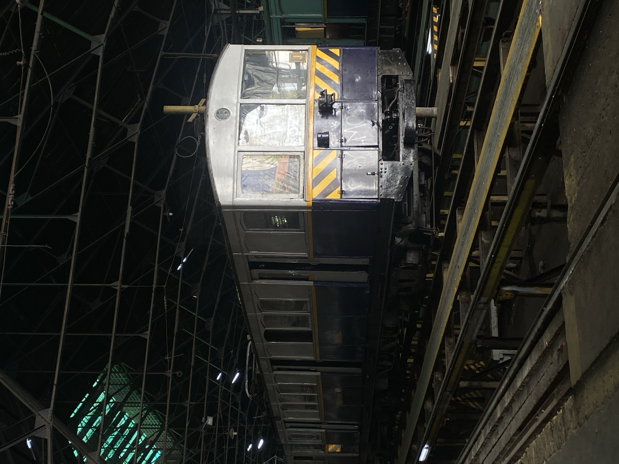 The image shows a subway car in what appears to be a maintenance facility. The car is predominantly gray and dark blue, with yellow and black diagonal stripes near the front. Graffiti is visible on the front of the car. The car is surrounded by a framework of metal beams and girders. The location is the Polvorín Workshop, a subway car maintenance facility in Buenos Aires, Argentina.