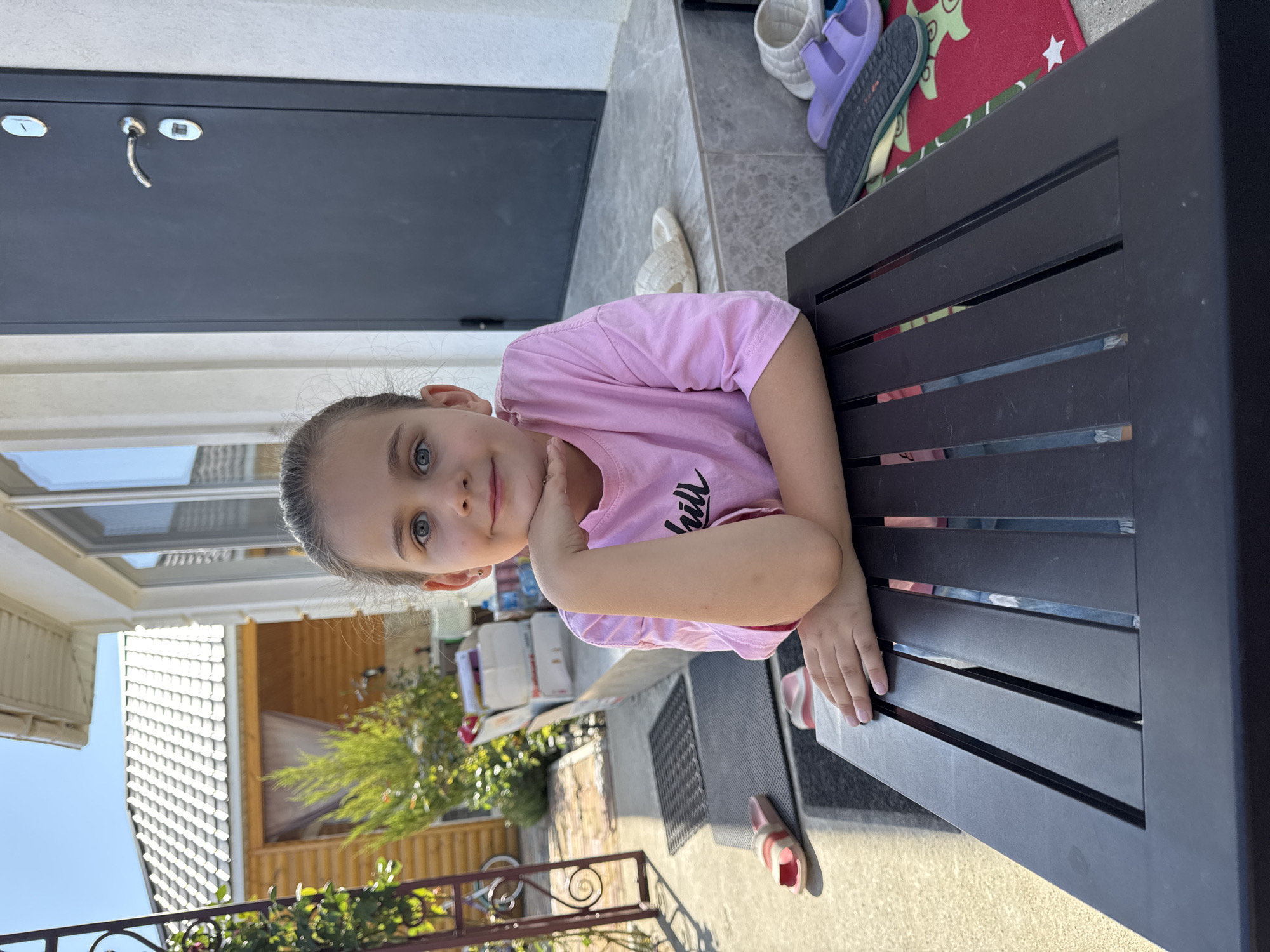 A young girl with brown hair pulled back is sitting at a dark brown, slatted outdoor table. She is wearing a pink t-shirt with the word "chill" printed on it. Her left arm is bent, and her hand is supporting her chin as she looks directly at the camera with a slight smile. She has blue eyes and fair skin. The background shows a house with a white facade and a dark door with a silver handle. To the left of the house, there is a wooden structure with a grey roof, and some greenery. Several pairs of sandals are visible on the ground and steps of the house. The scene appears to be outdoors on a sunny day, likely in a residential area.