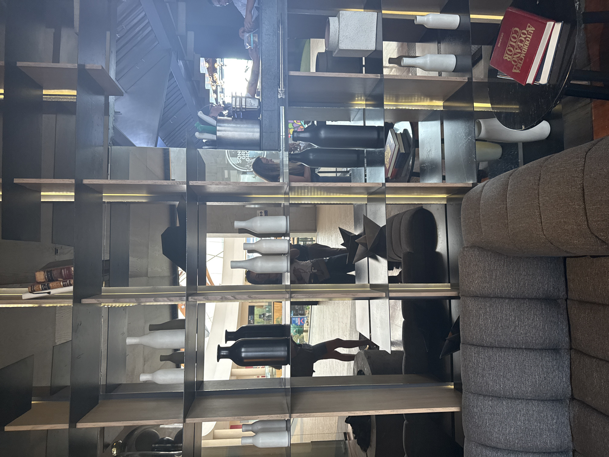 The image shows the interior of a commercial space, likely a restaurant or bar, in the city of Puebla, Mexico. A large, open shelving unit displays a variety of decorative objects, including white and black bottles of different shapes, books, and geometric sculptures. The shelves are illuminated by recessed lighting, creating a warm glow. The unit is positioned in front of a mirror, which reflects the interior of the space, including other patrons and staff. A grey sectional couch is situated near the shelving unit. The reflection in the mirror reveals a brightly lit space with other commercial establishments visible in the background.