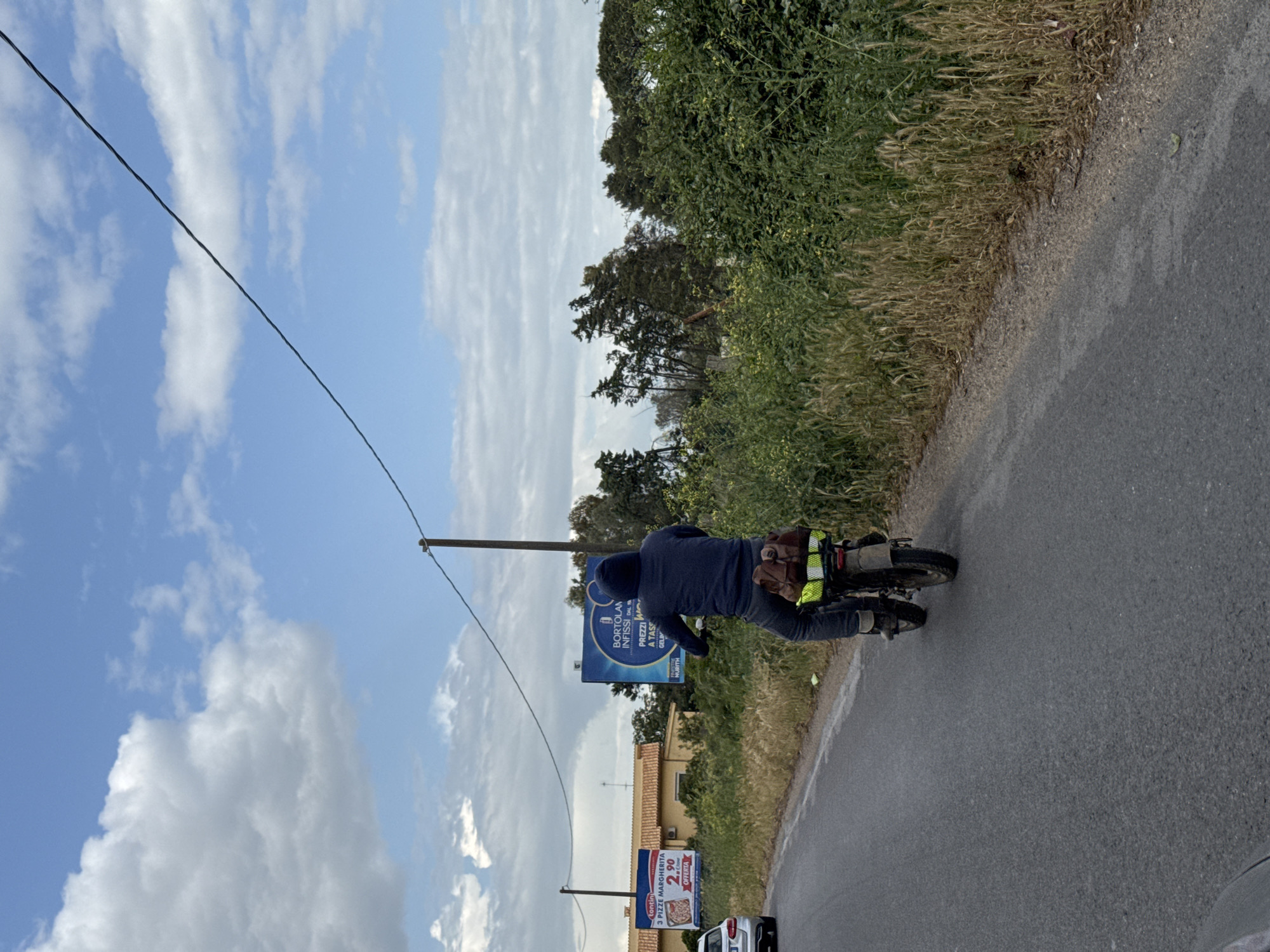 The image, taken in Nettuno, Italy, shows a person riding a motorized bicycle or small moped along an asphalt road on a clear, sunny day. The sky is bright blue with scattered white clouds. The person is viewed from behind, wearing a dark blue hooded sweatshirt, dark pants, and a high-visibility yellow vest over a brown bag or satchel. The bicycle/moped has gear attached to its rear.
To the right of the road, there's a verge covered in green foliage and dry grasses, with taller trees and bushes in the background. A utility pole with overhead wires stands on the left side of the road, running parallel to the direction of travel.
Two billboards are visible on the left side of the road. The upper billboard, partially obscured by the utility pole, advertises "BORTOLAN INFISSI DALA" (Bortolan Frames/Fixtures from) with "PREZZI A TASC" (prices you can pocket) and "GELIN," indicating a business selling building components. The lower billboard promotes "3 PIZZE MARGHERITA" for "2.90" as an "OFFERTA" (offer), with the brand names "tontin" and "NURITH" also visible. A small part of a white car is visible in the bottom left corner. The scene depicts a quiet, semi-rural setting during daylight hours.