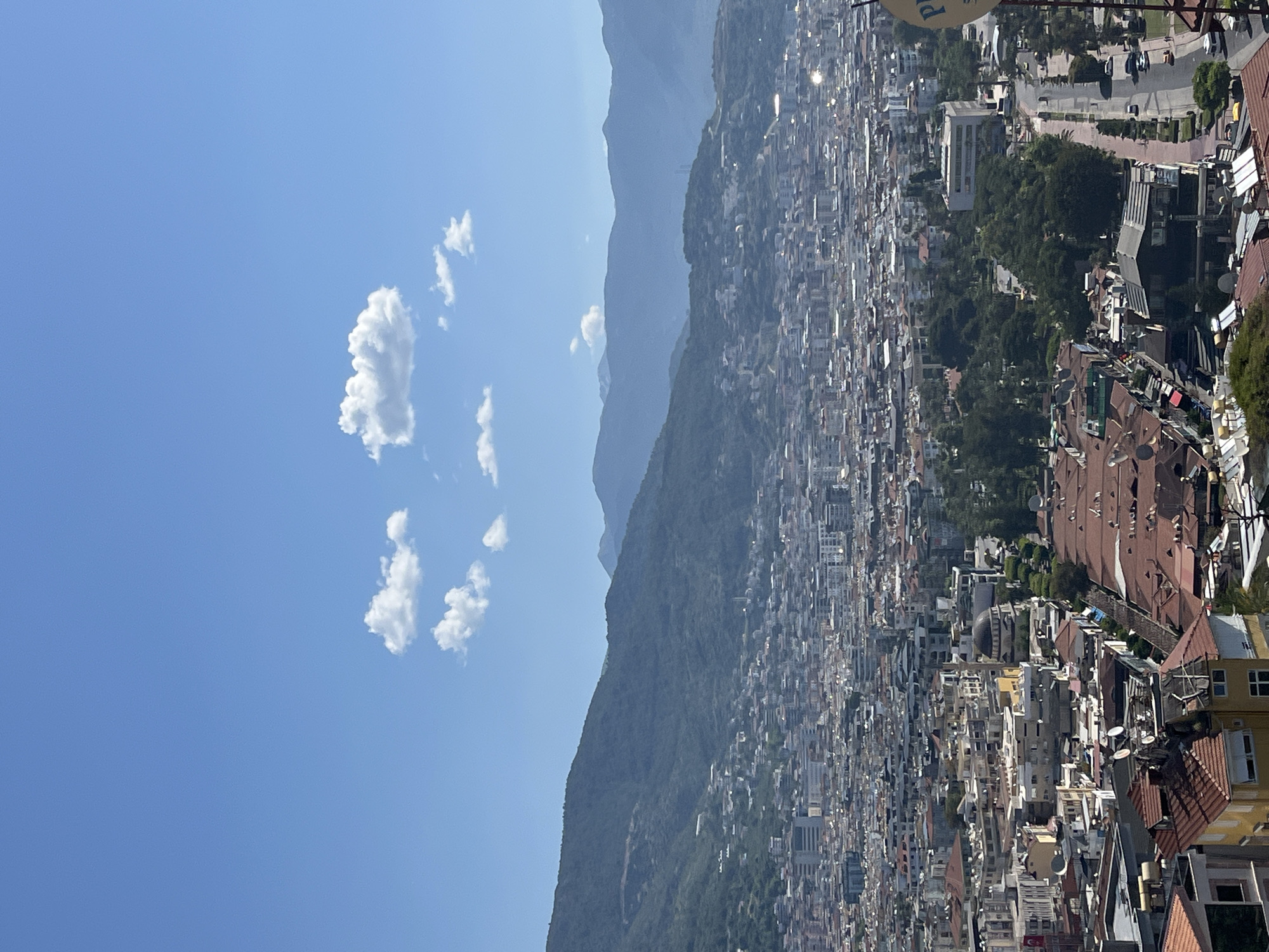 The image is a vertically oriented, high-angle shot of Alanya, Turkey. The scene is dominated by a dense urban landscape sprawling from the base of a mountain range toward the coast.
**Environment:**
The sky is a clear, bright blue with a few scattered cumulus clouds. The mountains are hazy and recede into the distance. The city is built on a mix of flat and sloped terrain.
**Objects:**
- A vast number of buildings, mostly low-rise and densely packed, are seen. The architecture is varied, with a mix of white, tan, and red-tiled roofs.
- Some taller buildings stand out amidst the smaller structures.
- Trees and greenery are interspersed throughout the urban fabric, particularly along the slopes of the mountains.
- A glimpse of the coast is visible in the far distance.
- Satellite dishes are visible on many rooftops.
- A sign partially visible in the top right, possibly of a restaurant or cafe.
**Location:**
The image captures a panoramic view of Alanya, Turkey. The city is known for its location on the Mediterranean coast, nestled at the foot of the Taurus Mountains. The dense urban development stretching from the mountains toward the coast is characteristic of the region.