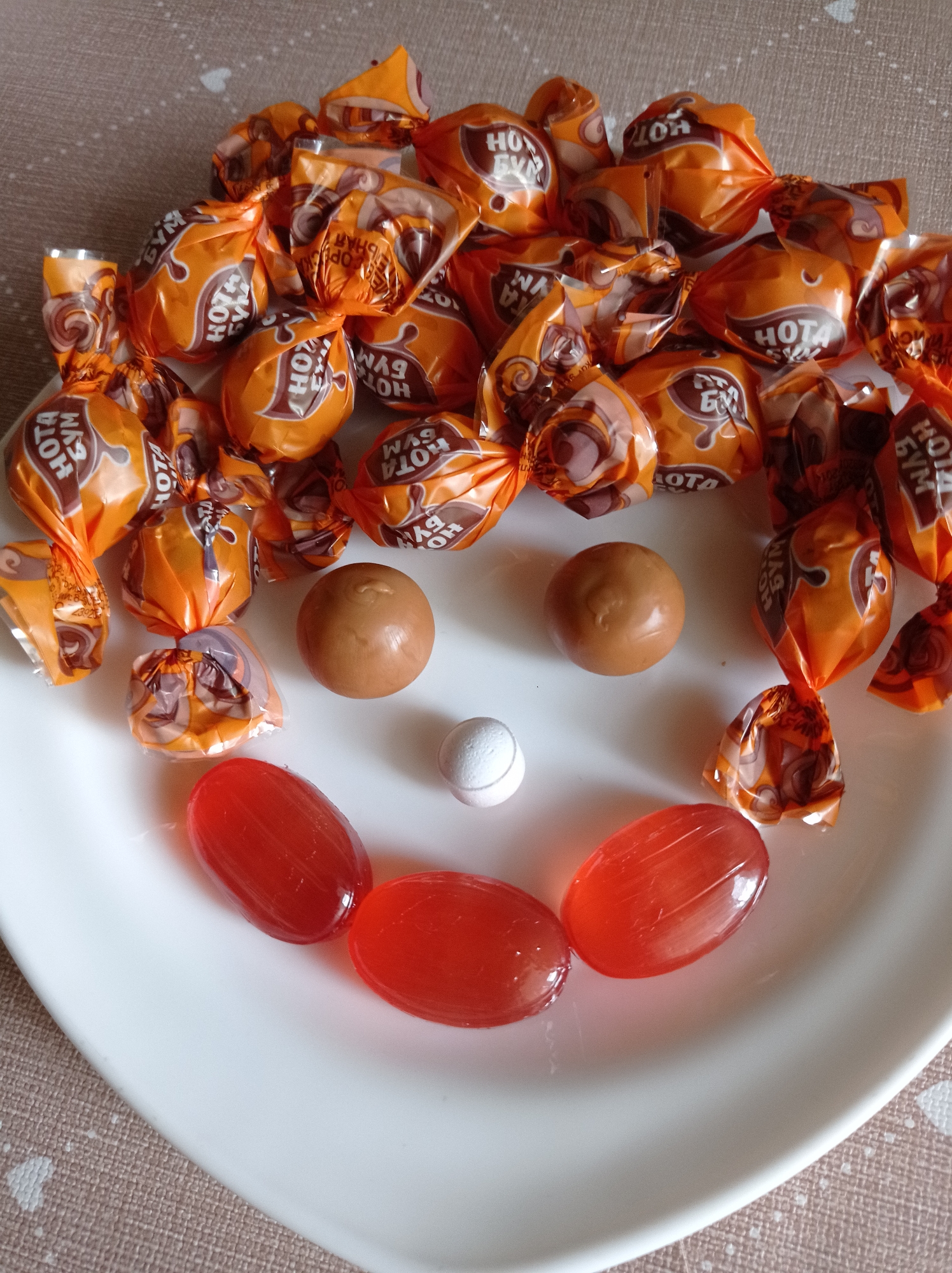 The image shows a white plate arranged with candies to resemble a smiley face. The 'hair' is made of wrapped "Hota Бум" candies, which are orange with brown lettering. The eyes are two unwrapped, caramel-colored round candies. The nose is a single, small, white, round candy. The mouth is formed by three red, oval-shaped hard candies. The plate sits on a beige surface with a geometric pattern. No specific location within Degtyarsk, Russia, is discernible from the image.