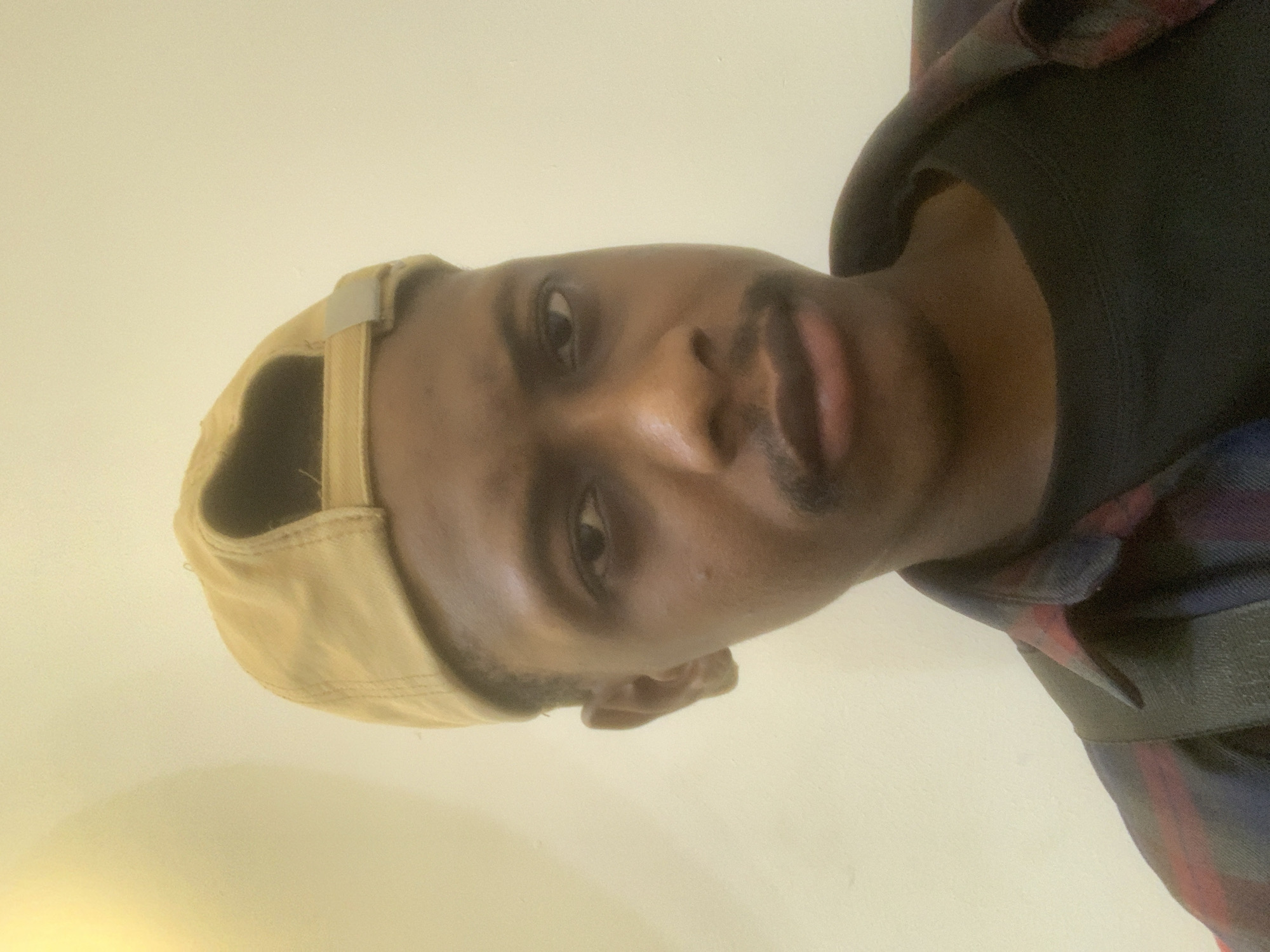 A man is featured in a close-up portrait, oriented vertically. He has dark skin and short facial hair, including a mustache and chin beard. He looks directly into the camera with a neutral expression. He is wearing a light tan or beige baseball cap backward, a dark t-shirt, and a plaid shirt with patterns of red, blue, and black over it. A dark strap, possibly from a bag, is visible over his right shoulder. The background consists of a plain, light-colored wall, suggesting an indoor setting. The lighting is soft and ambient. No visible text is present in the image. The location context is Animashawun, Nigeria.