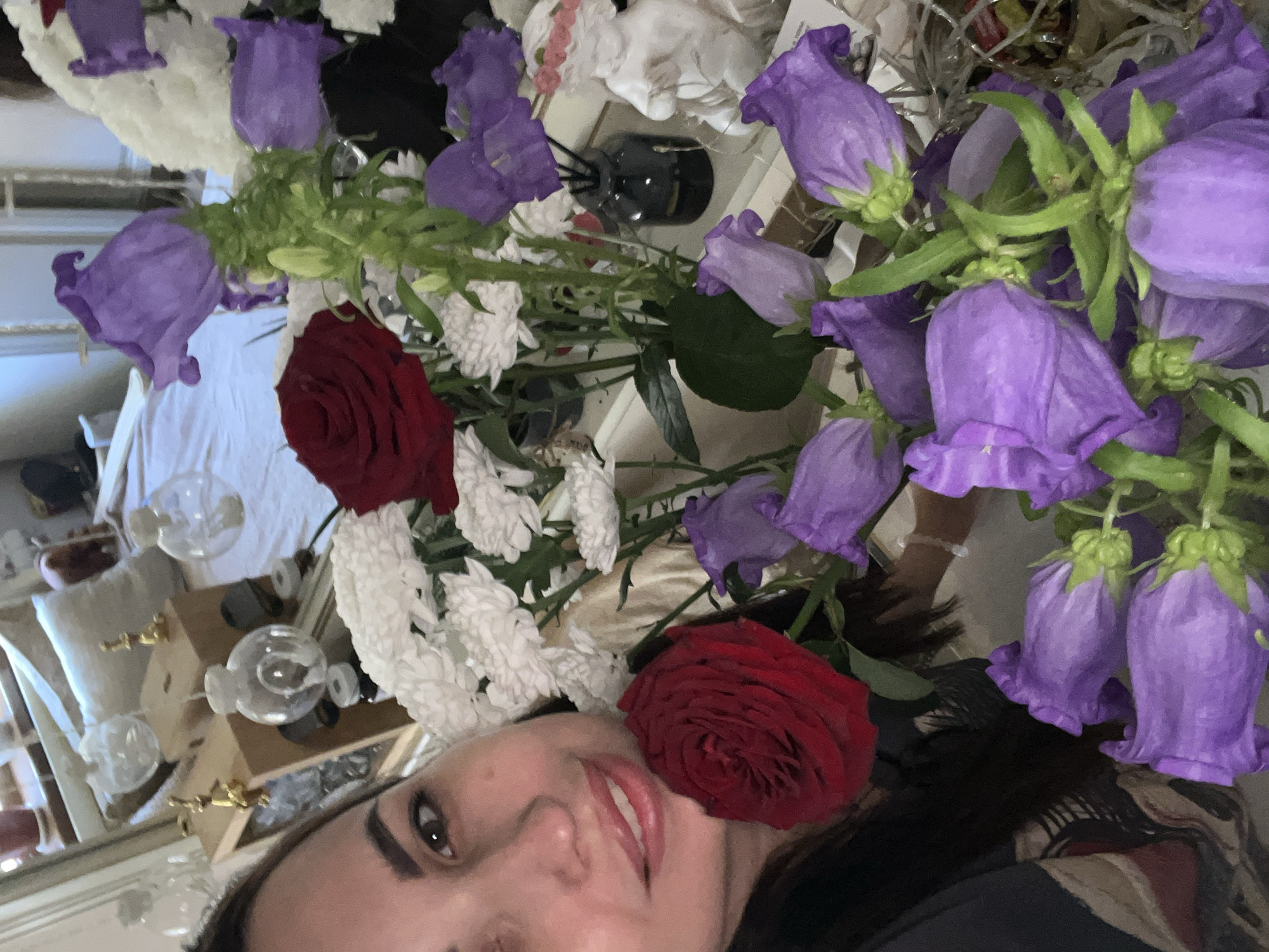 A woman with dark hair and brown eyes is in the foreground of the image, smiling at the camera. She is holding a bouquet of red roses and purple bellflowers. In the background, there are several decorative objects, including glass spheres and figurines, placed on a table. The scene appears to be indoors, possibly in a home.