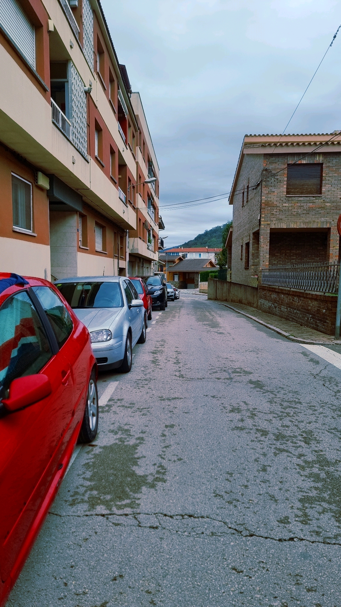 The image depicts a street scene in Sant Joan Les Fonts, Spain. On the left side of the street is a multi-story building with orange and beige facades, featuring balconies with metal railings and white latticework. Several cars are parked along the curb in front of the building. The closest car is red and viewed from the side. Behind it are a silver Volkswagen Golf, a red car, a black car, and several other vehicles further down the street. The street itself is asphalt, wet and appearing somewhat patchy, with visible cracks. On the right side of the street, there is a brick building with a tiled roof and a dark-colored shuttered window. A black metal fence runs along the property line of the brick building. In the background, more houses with red roofs and trees can be seen, with a hill or mountain rising behind them. The sky is overcast and appears cloudy. The overall impression is of a residential street, possibly after rain.
