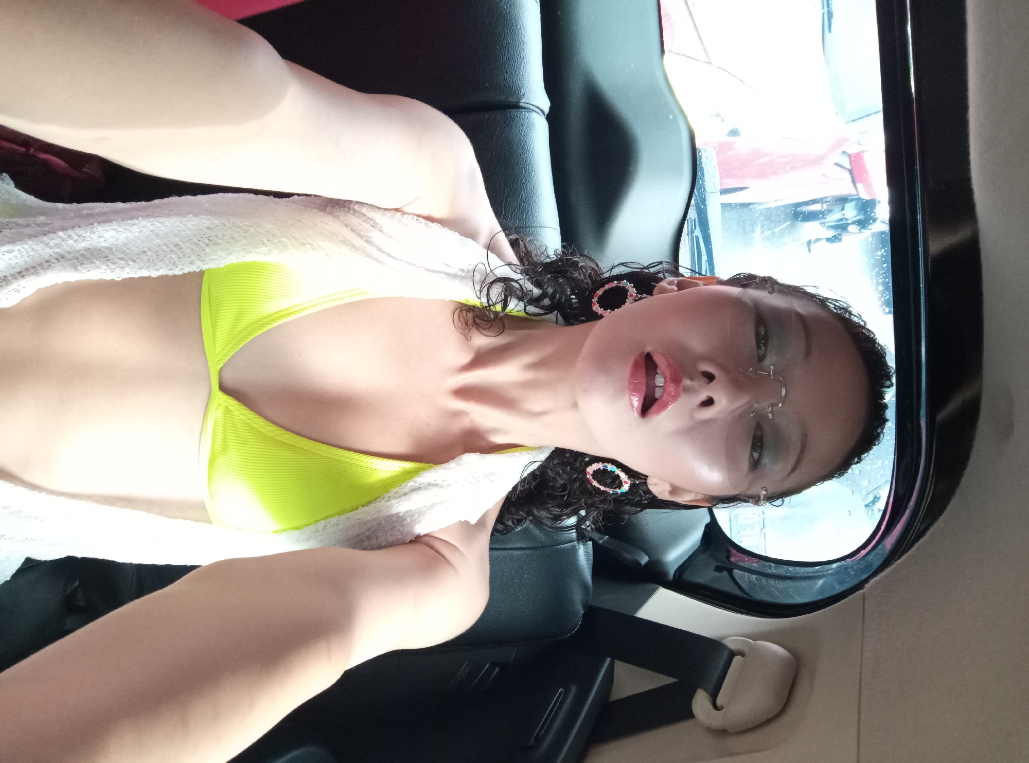 A woman is seated in a car. She is wearing a yellow bikini top and a white towel is draped over her shoulders. She has dark curly hair and is wearing earrings and glasses. The car window shows a blurry view of a red vehicle and other indistinct objects. Rain or water is visible on the window. The car's interior has black seats and a seatbelt. The location is identified as North Kuta, Indonesia.