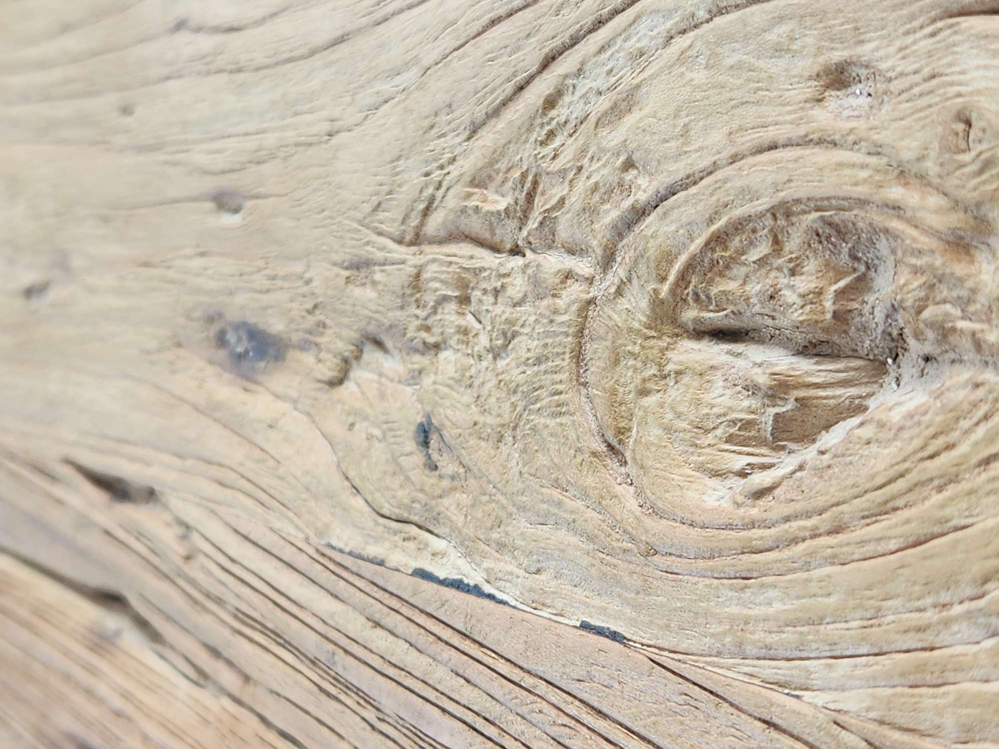 This is a close-up, high-angle shot of a weathered, light-colored wooden surface, likely a piece of driftwood or an aged wooden plank. The image focuses on the intricate grain patterns and textures of the wood. Prominent features include a large, circular grain pattern resembling an eye or a knothole, with concentric rings and rough, textured inner surfaces. Parallel lines of the wood grain run diagonally across the lower portion of the frame, creating a sense of depth and direction. Darker specks and lines are visible within the lighter wood, adding to its aged appearance. The lighting appears to be natural and diffused, highlighting the three-dimensional qualities of the wood's surface, with shadows accentuating the ridges and hollows. There are no people, animals, or any discernible scene or setting other than the wood itself. The image evokes a sense of natural beauty, age, and the passage of time, without any specific time of day, weather, or location cues beyond it being from Maitland, United States of America. No text is visible.