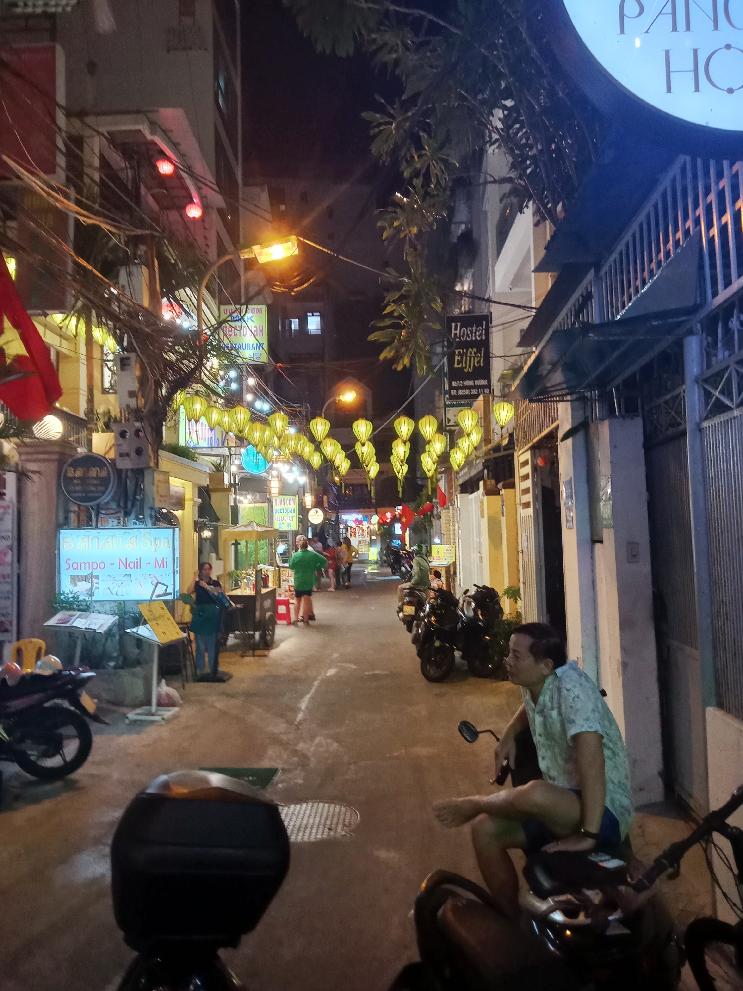 The image captures a narrow street in Nha Trang Ward, Vietnam, at night. The street is illuminated by strings of yellow lanterns and lights from business signs. Several motorbikes are parked along the street. A man sits barefoot on a parked motorbike in the foreground. Further down the street, a woman stands near a food stall. Other people are walking in the distance. Signs for businesses, including a spa and restaurants, are visible in both Vietnamese and English, and Korean. The "Hostel Eiffel" sign and overhead wires add to the urban atmosphere.