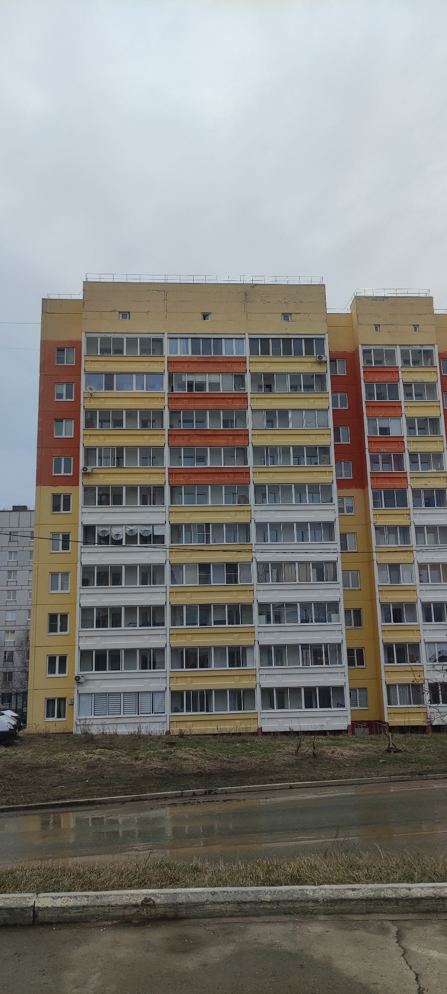 This image depicts a multi-story apartment building in Chaikovsky, Russia. The building is characterized by its alternating yellow, white, and orange facade. The windows are uniformly rectangular, and some have air conditioning units installed. The building has a flat roof with a metal railing. The foreground features a grassy verge and a wet, paved road. The sky is overcast, suggesting a dull or rainy day. Another, smaller apartment building can be seen in the background on the left.