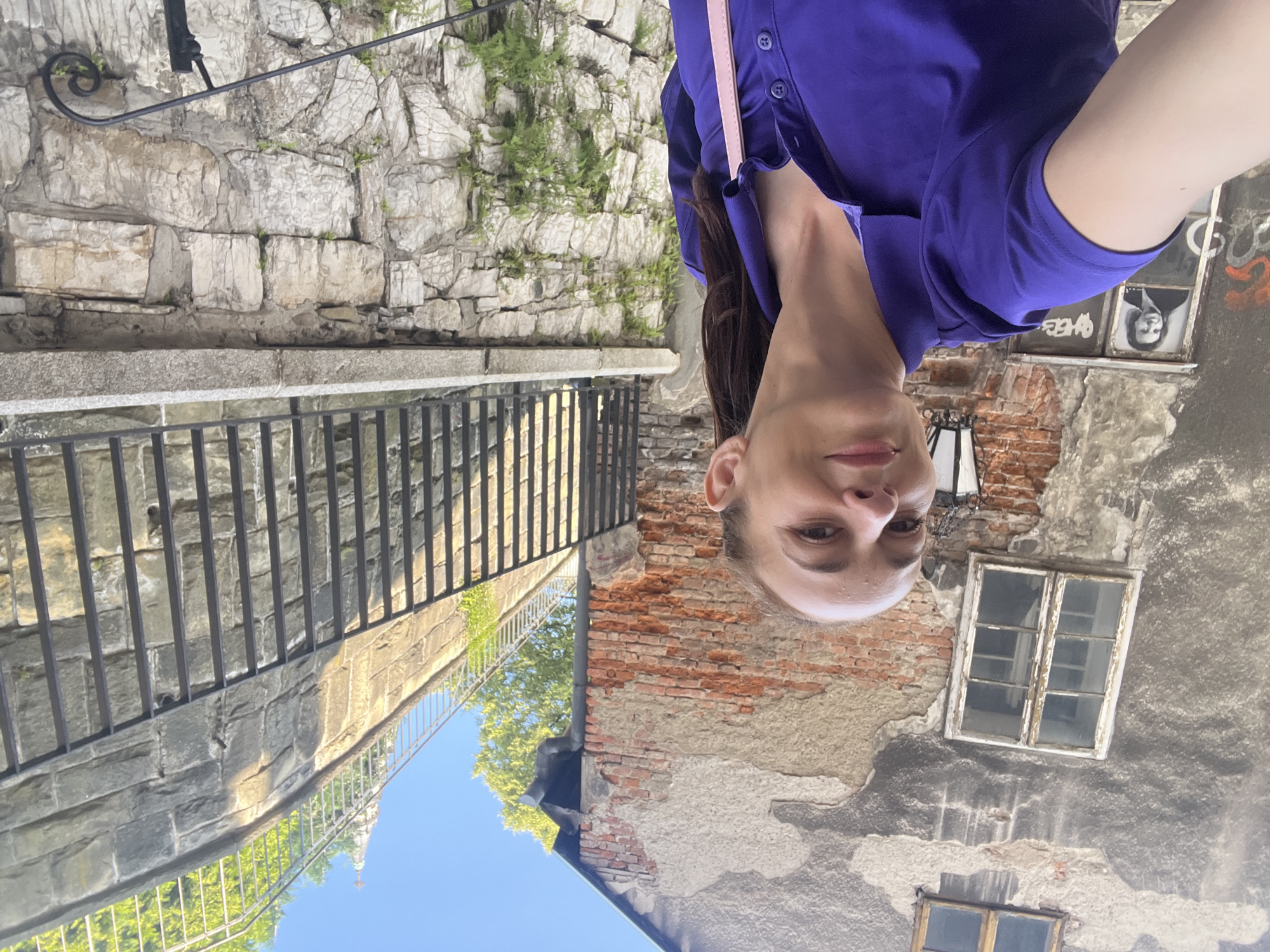 A woman is taking a selfie in an urban environment with old brick buildings and stone walls. She is wearing a purple polo shirt and has her hair pulled back. Behind her, there is a building with peeling plaster and exposed brick, two windows, and a street lamp. To the right of the building, there is a tall stone wall with a black metal fence and greenery. Further in the background, a steeple is visible against a clear blue sky. The woman is looking at the camera and smiling.