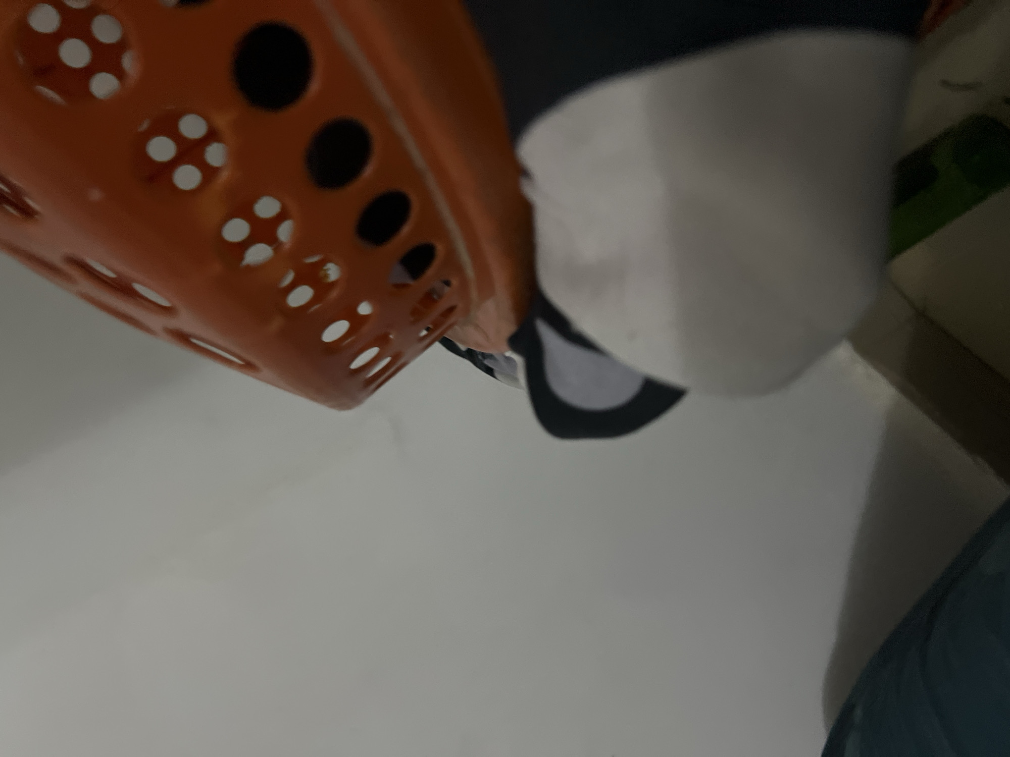 The image depicts an indoor scene, likely a residential setting. A portion of an orange plastic basket with circular perforations is visible at the top right, angled towards the viewer. Below the basket, a white object with a black edge is partially visible, suggesting it might be a stuffed toy or cushion. The background is a plain, light-colored wall. The lighting appears to be artificial, casting subtle shadows on the wall. There are no discernible landmarks or details that would allow for the identification of a specific location within Howayi, Nigeria. The image does not contain any identifiable individuals or actions.