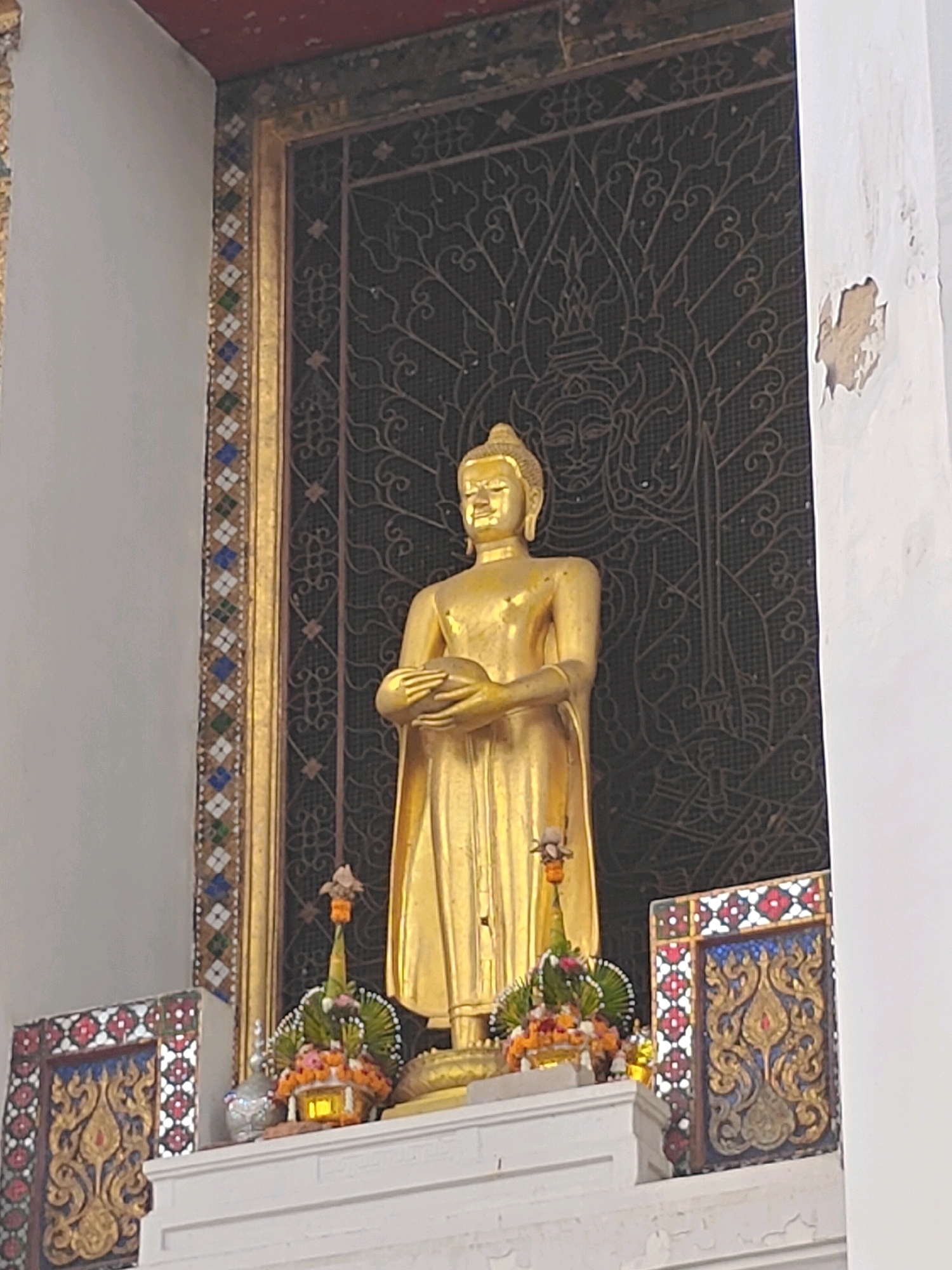 The image shows a golden Buddha statue standing in front of a dark, ornate metal screen. The screen features a stylized floral or tree pattern, with a subtle depiction of two faces integrated into the design. The Buddha statue is in a standing pose, with its hands cupped together in its lap, holding an alms bowl. It is draped in a golden robe.
To the left and right of the statue, there are decorative panels with intricate golden patterns and colored tile borders. In front of the statue, on a white ledge, are two small, pyramid-shaped floral offerings, adorned with small pink and orange flowers and green foliage. A small, illuminated shrine-like object is visible within one of these offerings.
The surrounding architecture includes a white wall with some peeling plaster on the right side, and a dark-colored border at the top, possibly a ceiling or roof edge. The overall setting appears to be a religious or cultural site. The image is shot from a slightly low angle, looking up at the statue. The lighting is somewhat diffused, creating soft highlights and shadows. The exact location within Phra Nakhon Si Ayutthaya City Municipality, Thailand cannot be definitively determined from the image alone, but the style of the Buddha statue, the ornate decorations, and the architectural elements are consistent with temples found in that region.