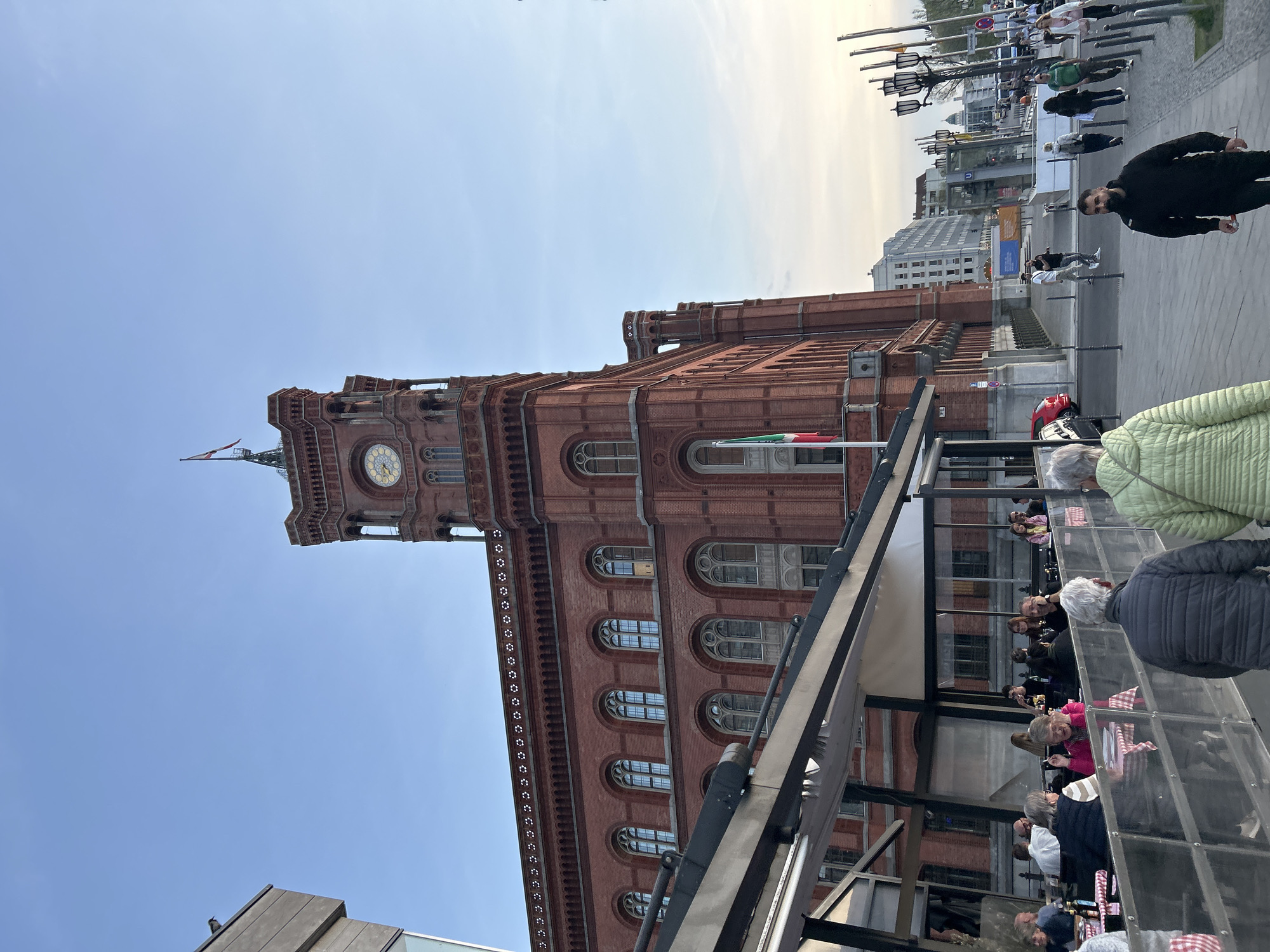 The image captures a view of the Rotes Rathaus (Red City Hall) in Berlin, Germany. The building, constructed with red bricks, stands prominently against a blue sky, partially obscured by the structure of what appears to be an outdoor dining area. People are seen seated at tables, some engaged in conversation. Others are standing and walking in the background, with a streetscape including lampposts and buildings visible. An Italian flag is flying from a flagpole attached to the building.