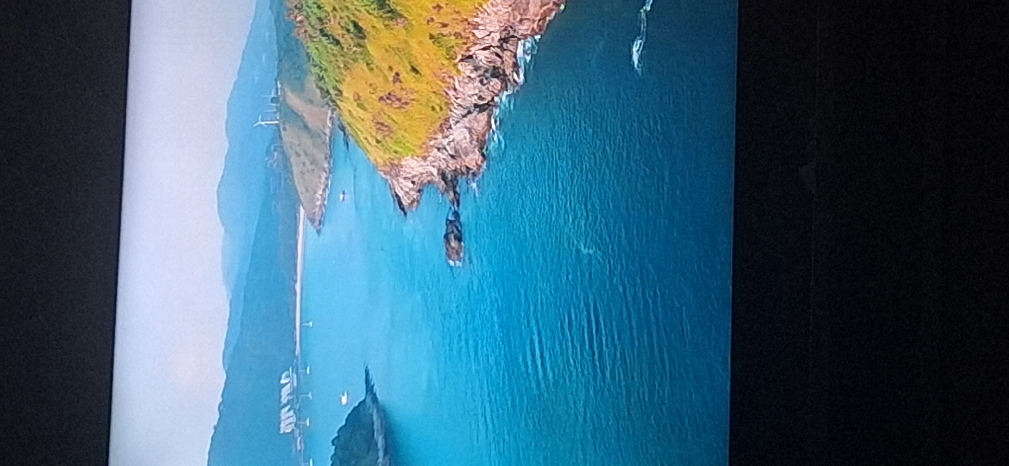 The image displays a serene coastal landscape. A tranquil body of blue water, likely an ocean or large bay, dominates the foreground and midground, with gentle ripples on its surface. Along the right side of the water, a rugged, rocky coastline is visible, covered in patches of green vegetation and golden-hued grass. Further inland, rolling hills rise, also sparsely covered with greenery and some trees. In the distance, hazy blue mountains form the backdrop. On the crest of one of the hills, several white wind turbines are clearly visible, suggesting a renewable energy source. In the far left of the midground, several white sailboats are anchored in the water near a sandy beach and more distant hills. The sky is a pale, overcast blue. This scene does not depict any part of the city of Bila Tserkva, Ukraine, as the geography is inconsistent with that location.