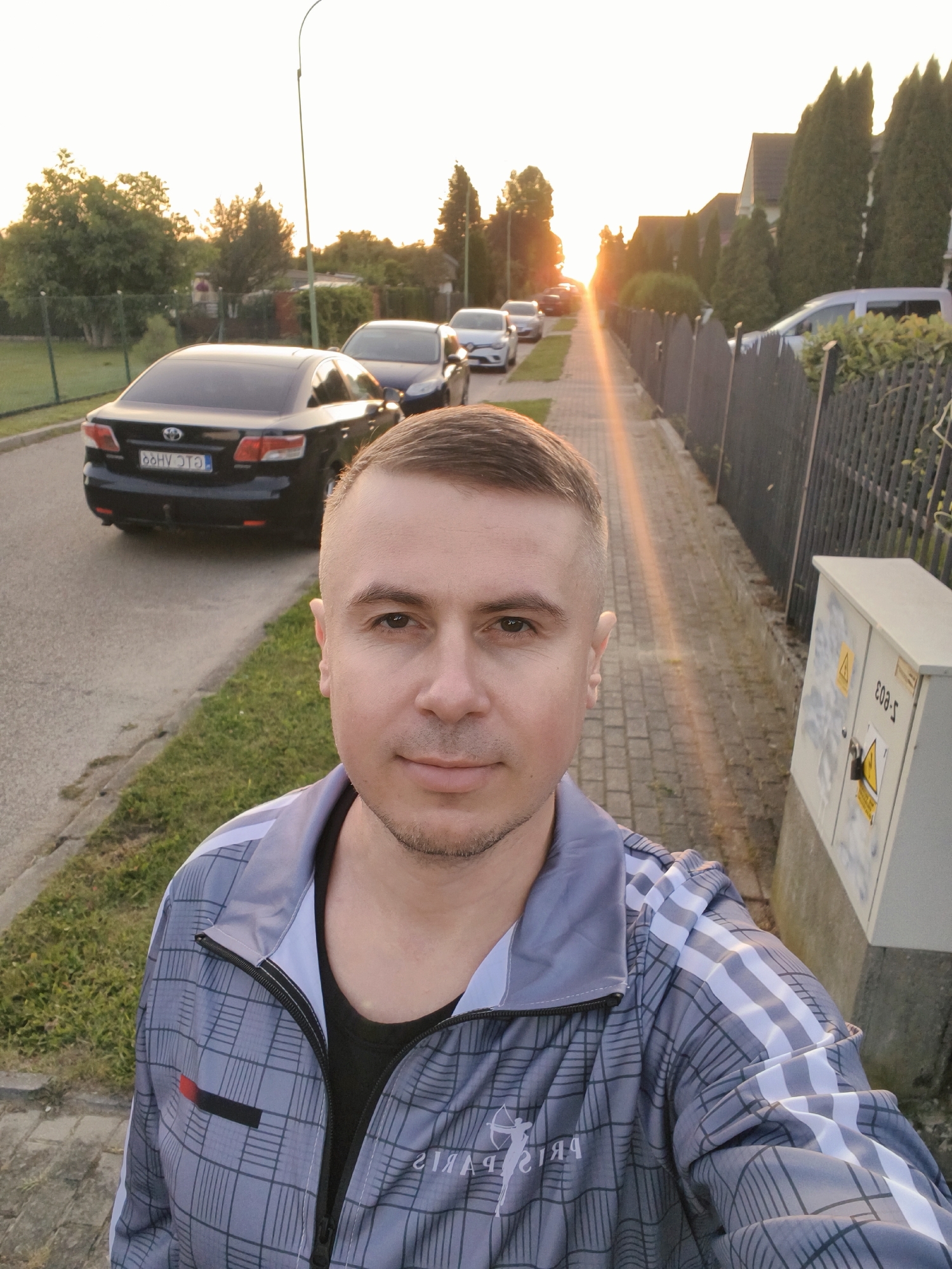 A man with short brown hair and stubble is taking a selfie outdoors, facing the camera. He is wearing a grey checkered jacket with white stripes on the sleeves and a black t-shirt underneath. The background shows a residential street with parked cars, houses with trees, and a sidewalk. The sun is setting, casting a warm glow and lens flare across the scene. On the right side of the image, a utility box is visible. The license plate on the black car in the foreground appears to be 66HVJ73. The overall environment suggests a quiet, suburban setting in Tczew, Poland, during a late afternoon or early evening.