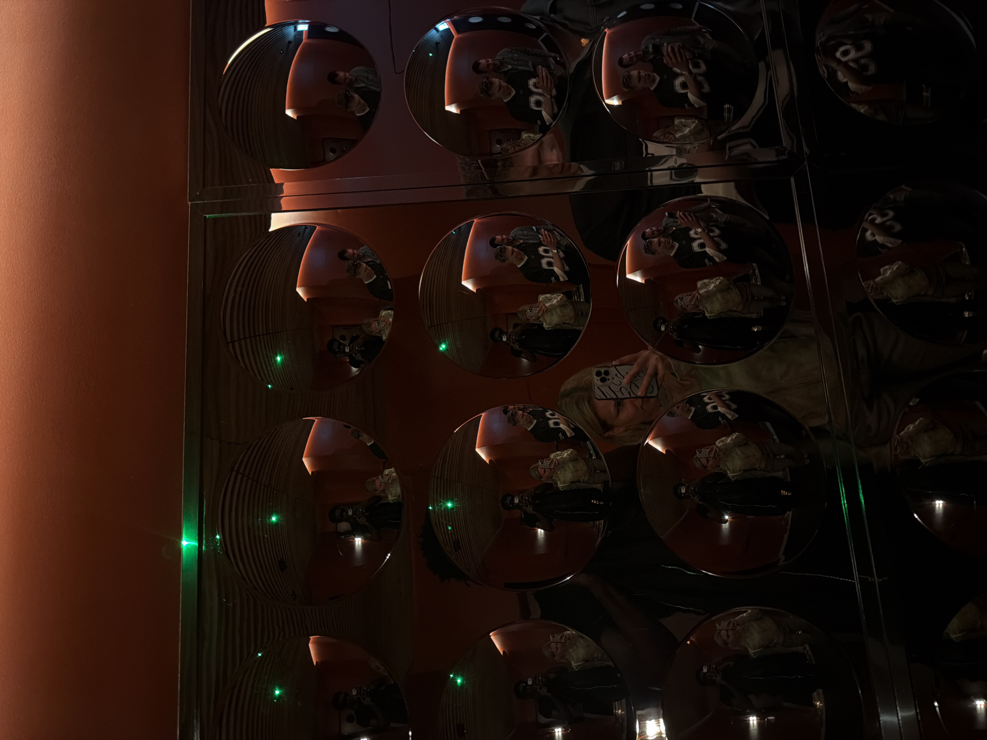 The image shows a wall covered in round mirrors, arranged in a grid pattern. The wall is next to a salmon-colored wall, reflecting green lights in some mirrors. Reflections of people and objects are visible in the mirrors, suggesting the location is inside a public space, possibly a restaurant or event venue, located in the city of London, United Kingdom. A person is visible holding a phone in their hand with the camera directed towards the mirrors.