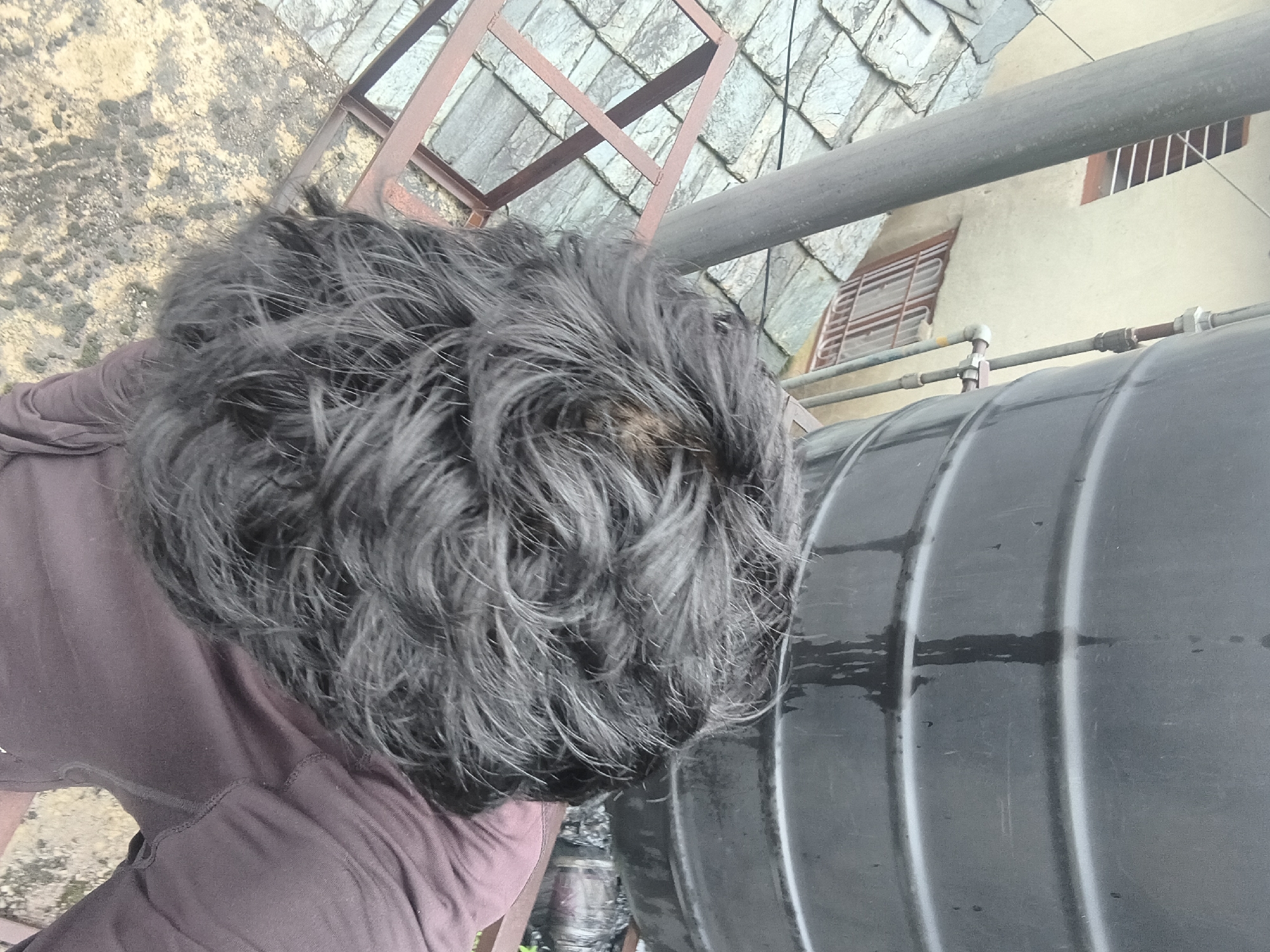 The image shows a close-up view of a person's head and shoulders, taken from above. The person has dark, messy hair. They are wearing a dark-colored garment. In the background, there is a large black water tank, a metal railing, and a sloped roof covered with grey tiles. Further in the background, a building with beige walls and barred windows can be seen. The environment appears to be an outdoor urban setting, possibly a rooftop or balcony. The image is grainy and appears to have been taken in natural light.