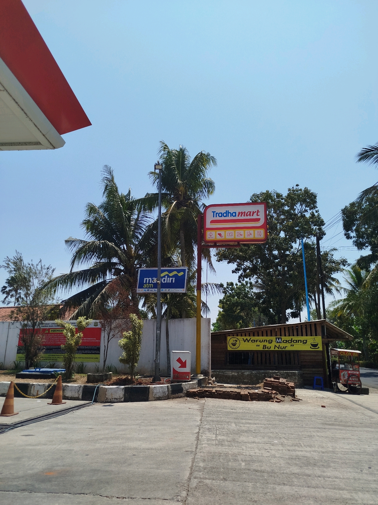 The image depicts a sunny outdoor scene in Bocor, Indonesia, likely at a gas station or rest stop. In the foreground, a paved surface shows some scattered bricks and concrete elements, along with two orange traffic cones and a yellow chain barrier. A prominent red and white canopy structure extends from the left side of the frame.
To the right, a small wooden structure with a thatched roof serves as a food stall, identified by a yellow banner reading "Warung Madang ” Bu Nur ”" and featuring a steaming cup logo. Next to it, a wheeled food cart is visible. Behind these structures, a white concrete wall is adorned with signs, including a blue sign for "mandiri atm" and a larger red and white sign for "Tradha mart". Palm trees and other foliage are scattered throughout the scene. The sky is a clear, bright blue. There are no people clearly visible in the image.