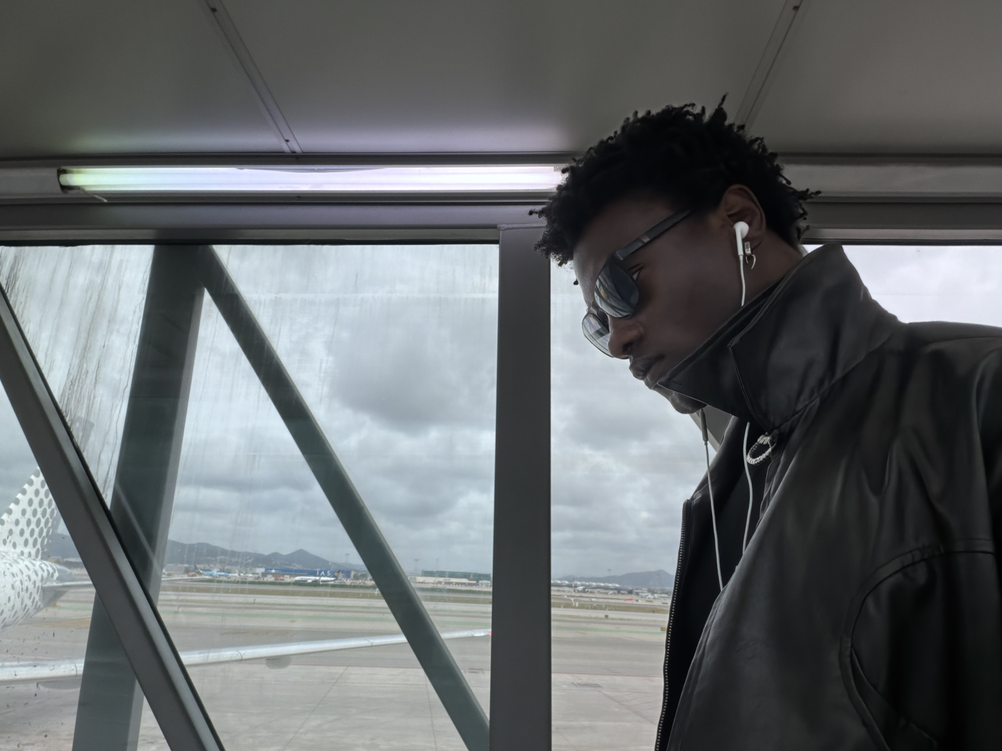 A man stands in a terminal jet bridge at Barcelona-El Prat Airport in El Prat de Llobregat, Spain. The man has short, dark hair and wears sunglasses, and wired headphones. He wears a black jacket. Through the jet bridge windows, the airport tarmac and distant hills are visible on a cloudy day. A plane is parked outside the window.