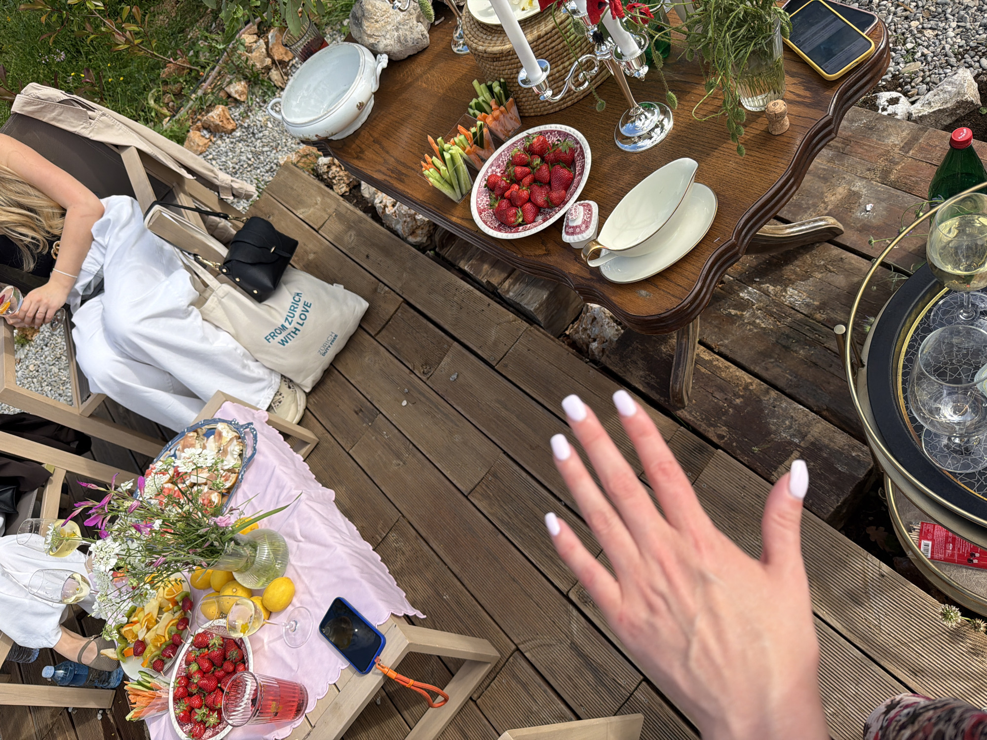 The image captures an outdoor setting on a wooden deck in Dobra Voda, Montenegro. Two tables are arranged for a meal. One table, covered with a pink cloth, has fruits, vegetables, and flowers. A second table holds strawberries, vegetables, a white ceramic dish, and decorative candles. Two women are present: one is sitting by the table with the pink cloth, and another person's hand with white nail polish is prominently displayed in the foreground. Objects like smartphones, bags, and drinks are scattered around the scene.