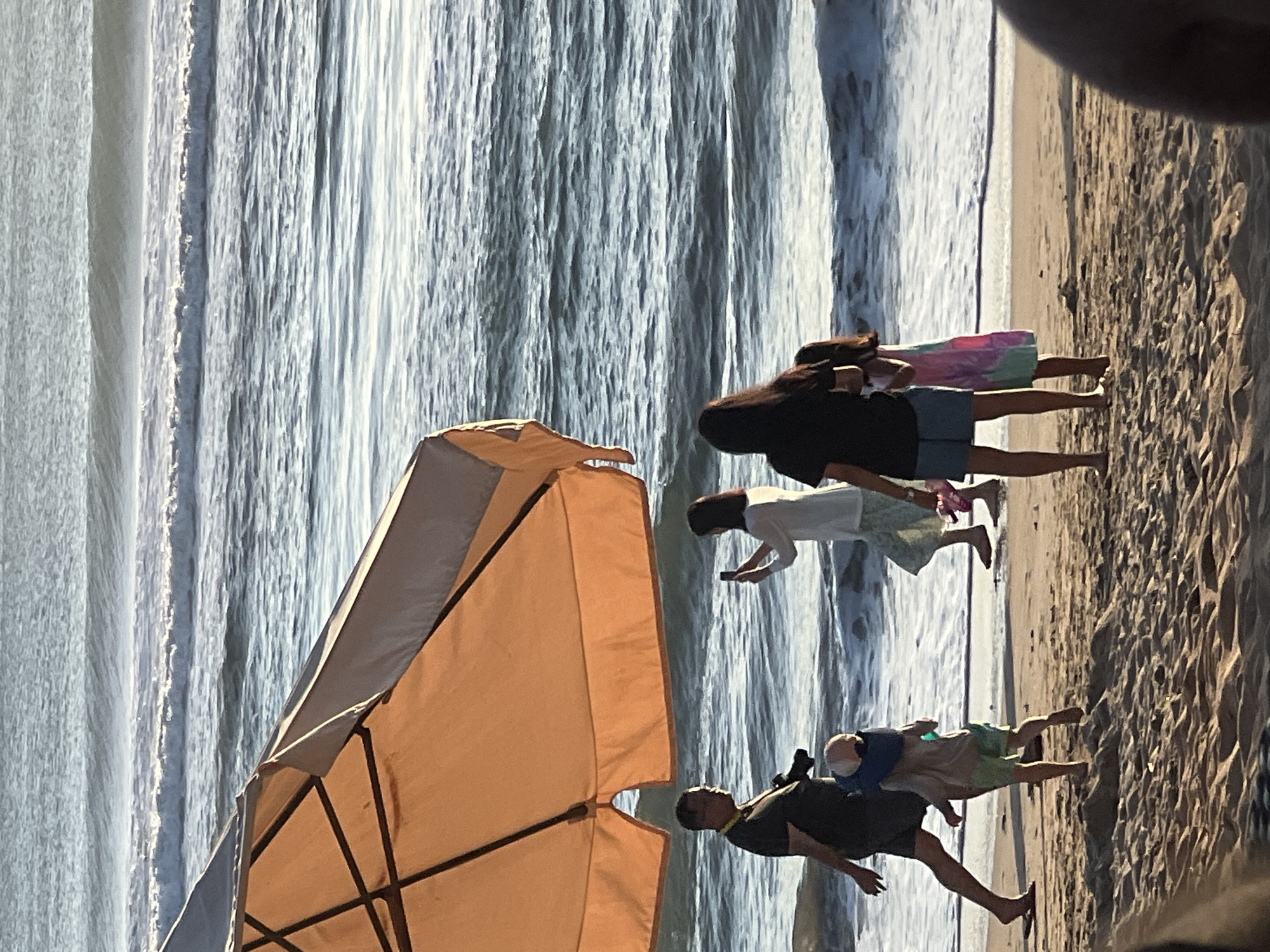 A group of people are on a beach in Kuta, Indonesia, with the ocean in the background. An orange beach umbrella is in the foreground, partially obscuring the view. A man with glasses is walking to the left, holding a camera. A child in a blue and green outfit is walking in front of him. A woman is taking a photo with her phone, and two young girls are standing next to her. The water has gentle waves, and the sand is light brown.