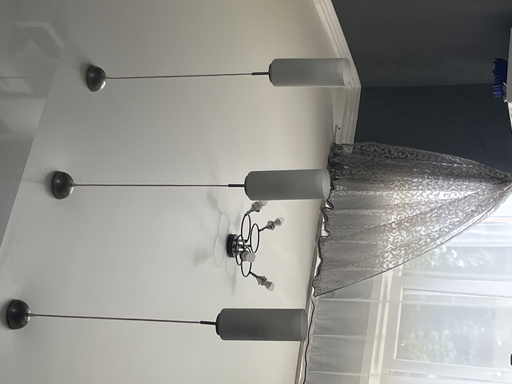 The image shows a room with three modern pendant lights hanging from the ceiling. The lights have cylindrical frosted glass shades and are suspended by thin wires. In the background, a decorative ceiling light fixture with multiple arms and light bulbs is visible. Sheer patterned curtains are drawn across a window on the left side of the frame, and a dark grey wall occupies the right side. A dark blue decorative bowl is on a surface in the lower right corner. The scene appears to be an interior residential space. No people or actions are depicted. The location of this scene within Saratov, Russia cannot be determined from the provided image.
