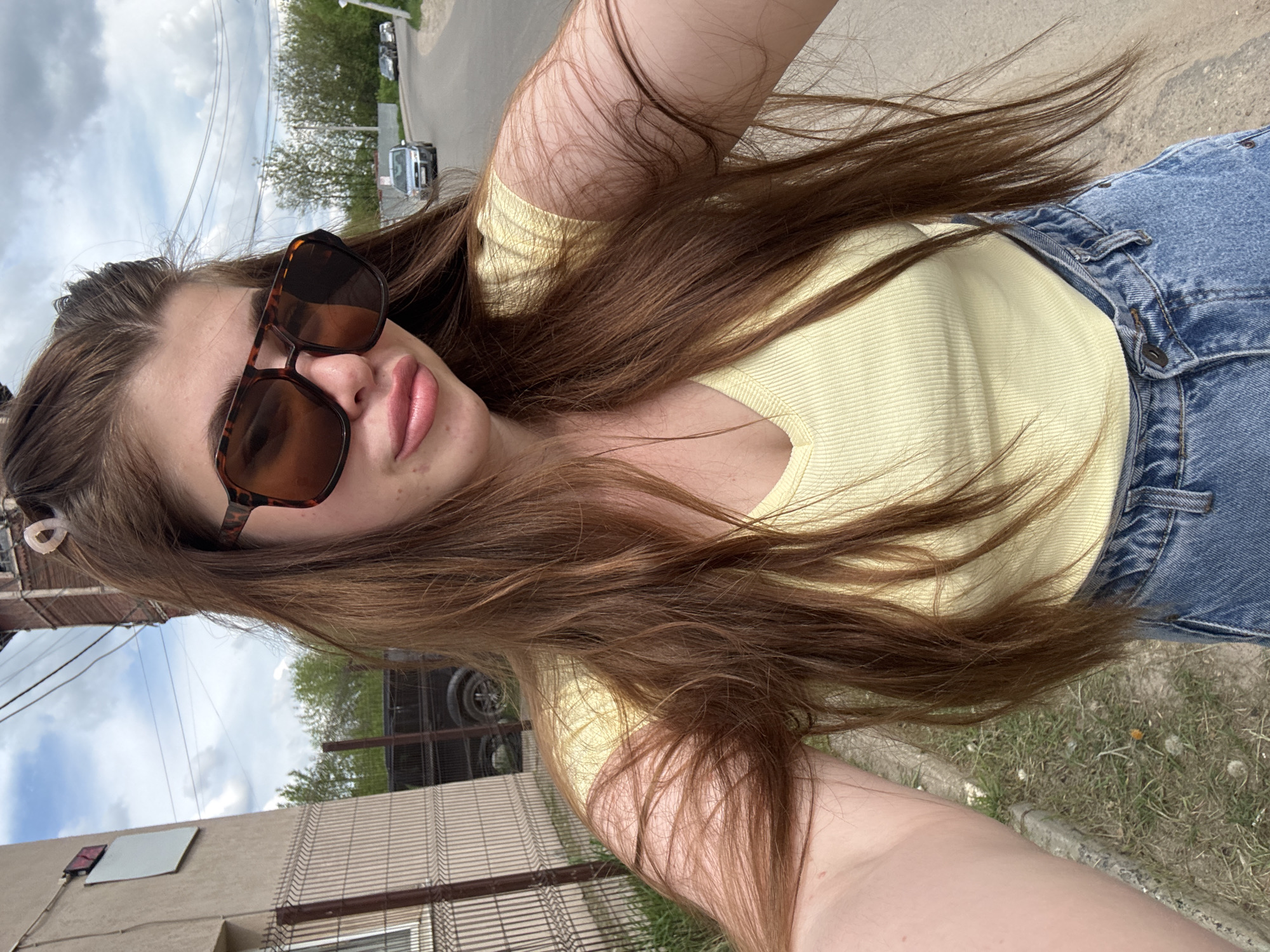 A selfie photograph was taken by a young adult in Посёлок комбината стройматериалов-1, Russia. The subject, wearing light-blue denim jeans, a yellow, short-sleeved top, and brown-framed sunglasses, is positioned at an oblique angle to the camera. The backdrop reveals a city street with buildings, a fence, and parked vehicles. The weather appears overcast, and natural light is diffused across the scene.