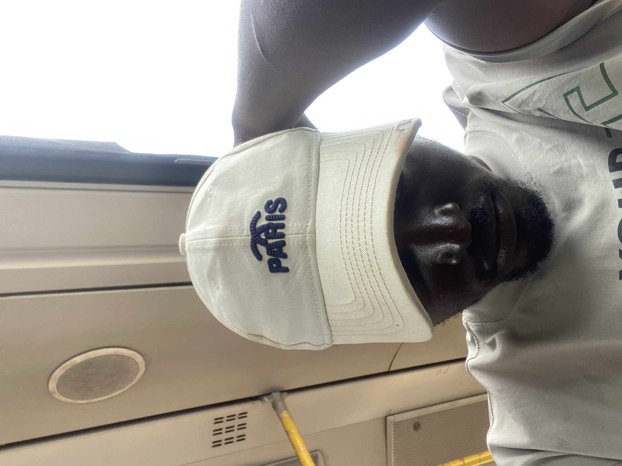 A person wearing a white baseball cap with "PARIS" embroidered on it in blue, and a grey t-shirt, is holding their right arm to the side of their head. They are looking down, with their face partially obscured by the cap. The background appears to be the interior of a vehicle, possibly a bus, with light filtering in from a window on the right side. A yellow pole is visible on the left side of the image, along with some ventilation grates and a circular speaker. The scene suggests the person is traveling.