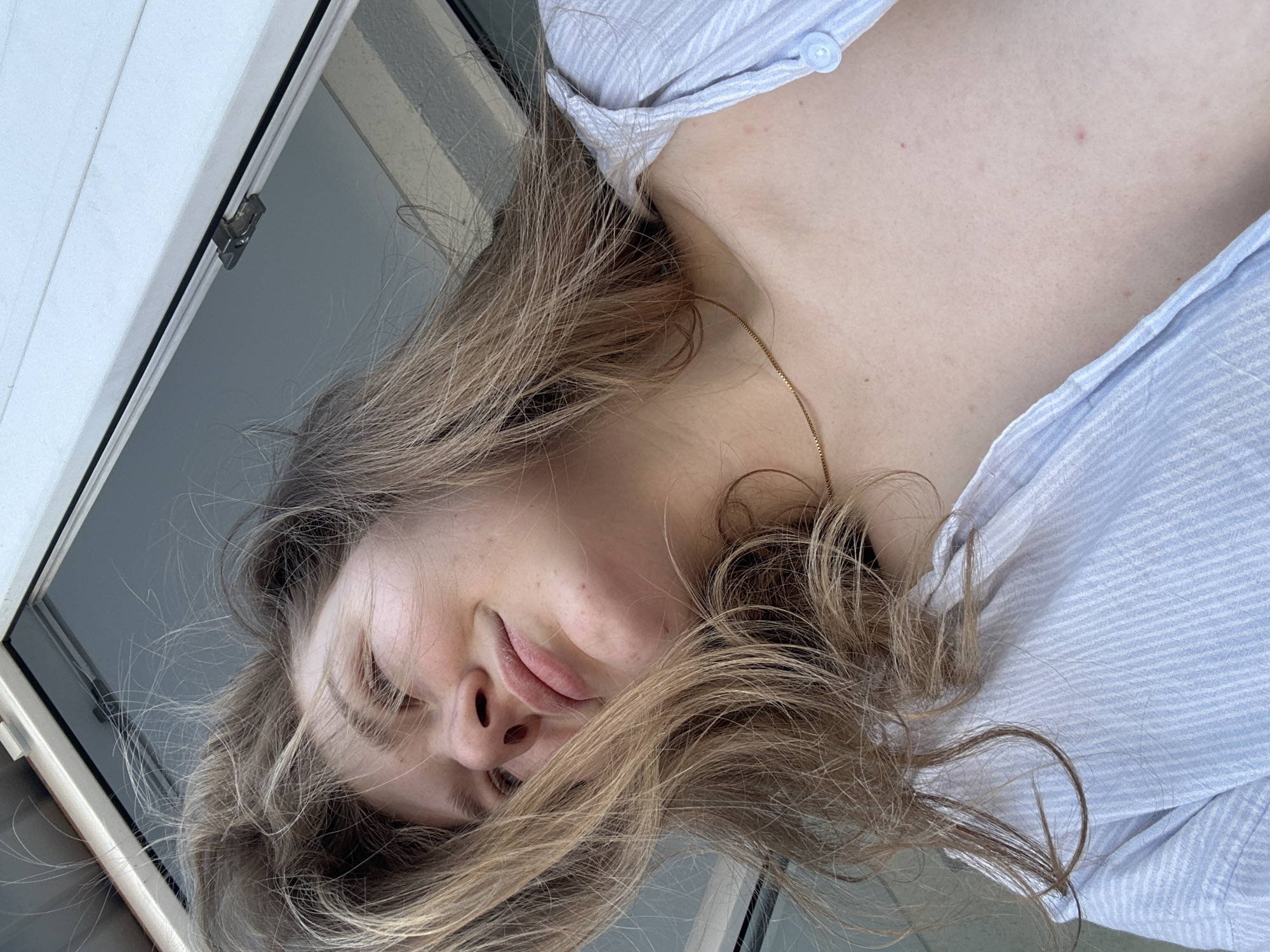 A woman with long blonde hair is lying down, wearing a light blue striped shirt. She has fair skin and a gold necklace. Her eyes are closed and she appears to be resting. She is near a window. No information is available to suggest the location is Kazan, Russia.