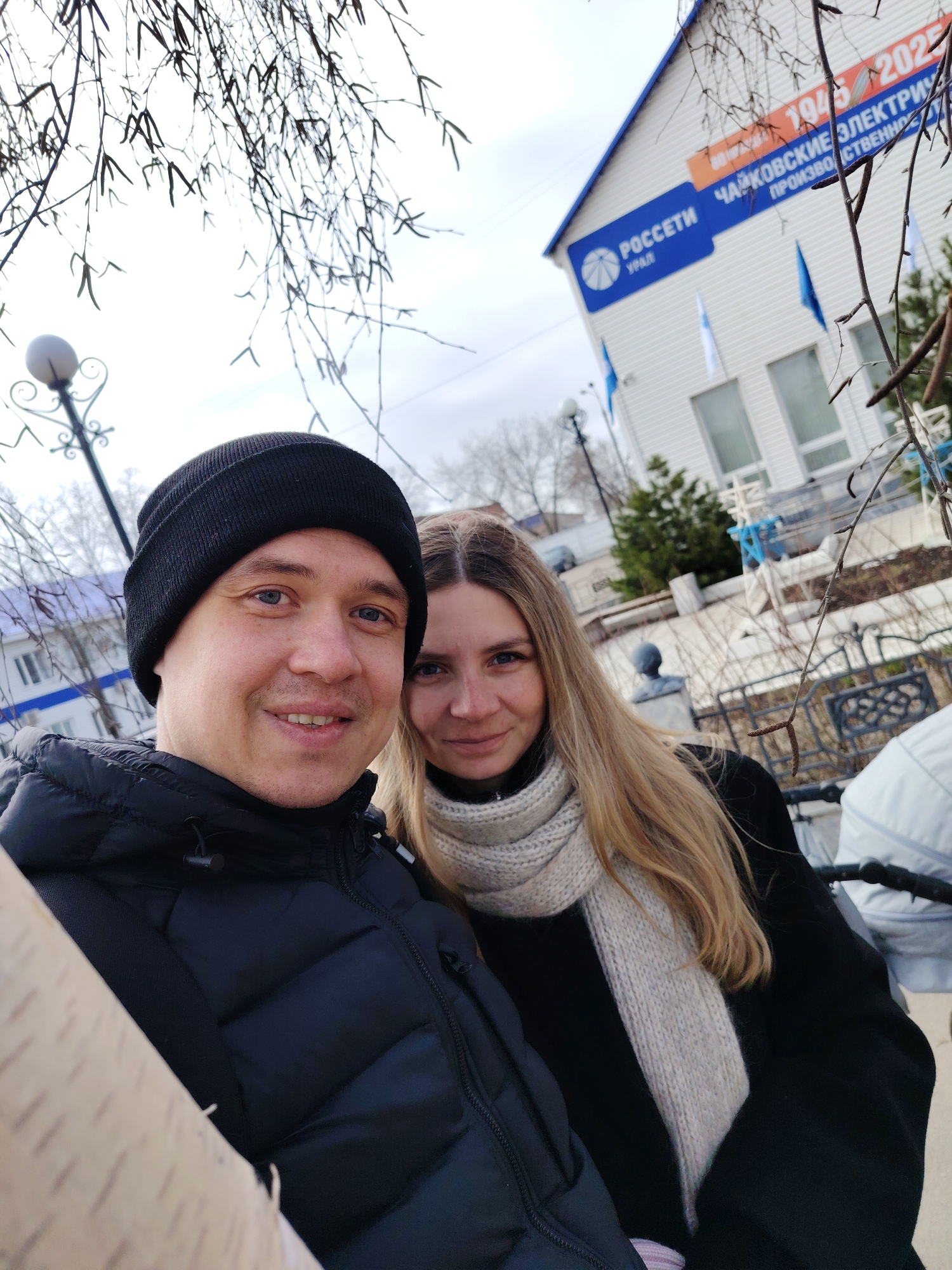 A selfie shows a man and a woman outdoors. The man wears a black beanie and a black puffer jacket. The woman is next to him, wearing a white scarf and a black coat. Behind them is a building with signage in Russian; the signs read "РОССЕТИ УРАЛ," "ЧАЙКОВСКИЕ ЭЛЕКТРИЧЕСКИЕ СЕТИ," and reference the years "1945-2025." The text indicates this is likely the Chaikovsky Electric Network Office in the city of Chaikovsky, Russia. A stroller is visible in the background near the right side. There are bare trees and street lamps in the background suggesting winter.