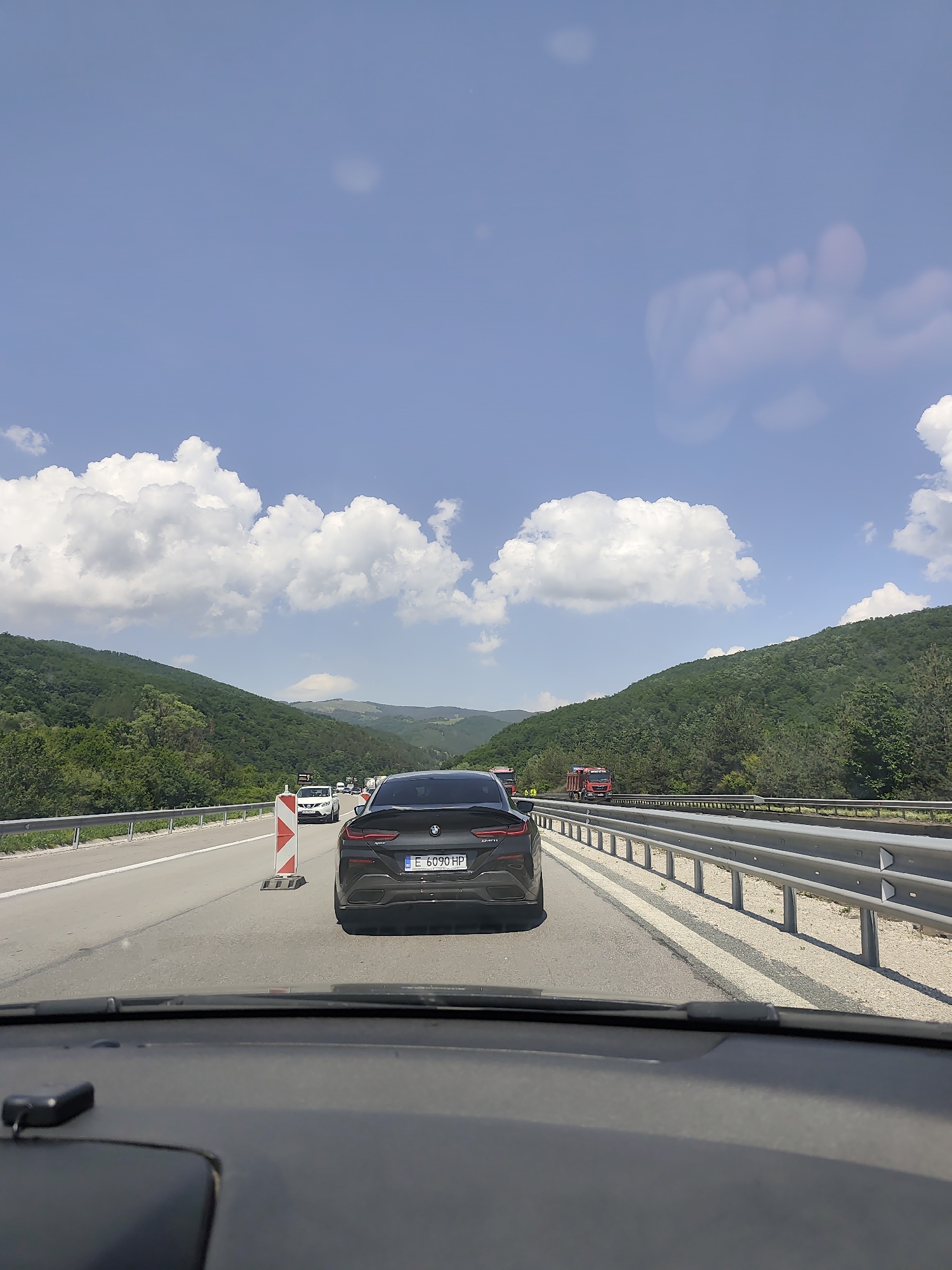 The image depicts a daytime scene on a multi-lane highway in a mountainous region under a clear blue sky with scattered white cumulus clouds. The perspective is from the interior of a vehicle, with the dashboard and windshield visible in the foreground.
In the midground, a dark-colored BMW, identified by its emblem and taillight design, is directly ahead. Its rear license plate reads "E 6090 HP". To the left of the BMW, a red and white striped traffic cone or barrier is positioned on the road. Further ahead in the same lane, a white SUV is visible. The highway has at least two lanes of travel in this direction, separated by clear road markings. A metal guardrail runs along the right side of the road, and another is partially visible on the left.
The background is dominated by lush green, tree-covered mountains or large hills rising steeply on both sides of the highway. On the opposite carriageway, several red dump trucks are visible. The sky above features several diffused, light-colored smudges, including a distinct, large, footprint-like shape in the upper right quadrant, likely reflections or debris on the camera lens or vehicle windshield. The overall environment suggests a rural or semi-rural setting within Unknown, Unknown, Bulgaria.