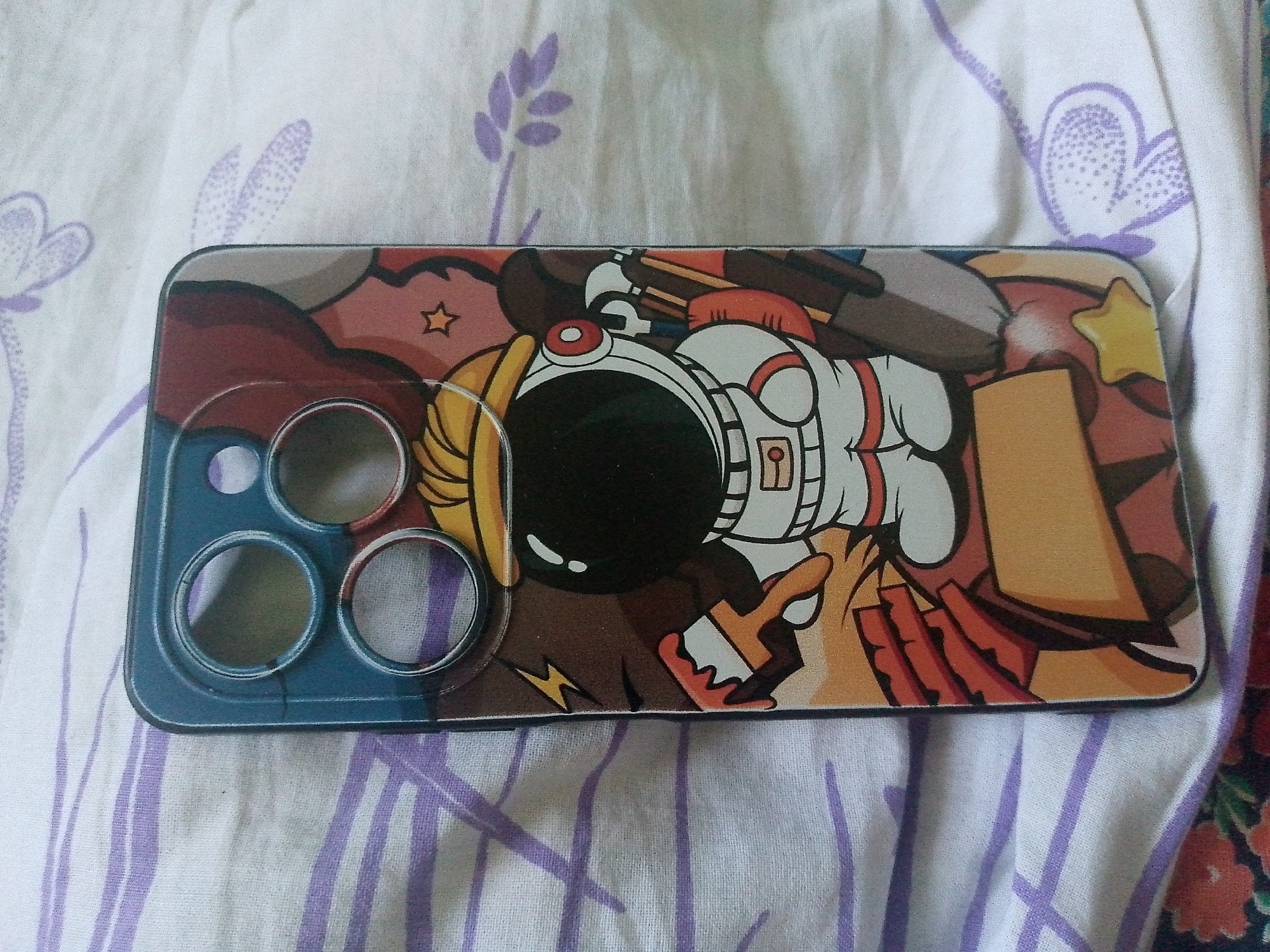 The image shows a smartphone case with an astronaut cartoon design. The case has a blue camera cutout area on the left side with three circular openings. The design features a cartoon astronaut with a white suit, yellow accents, and a black visor. The background is a mix of abstract shapes and colors, including brown, red, and orange. The case is lying on a white fabric with purple floral patterns and vertical stripes. The image does not contain any elements related to the city of Bukhara, Uzbekistan.