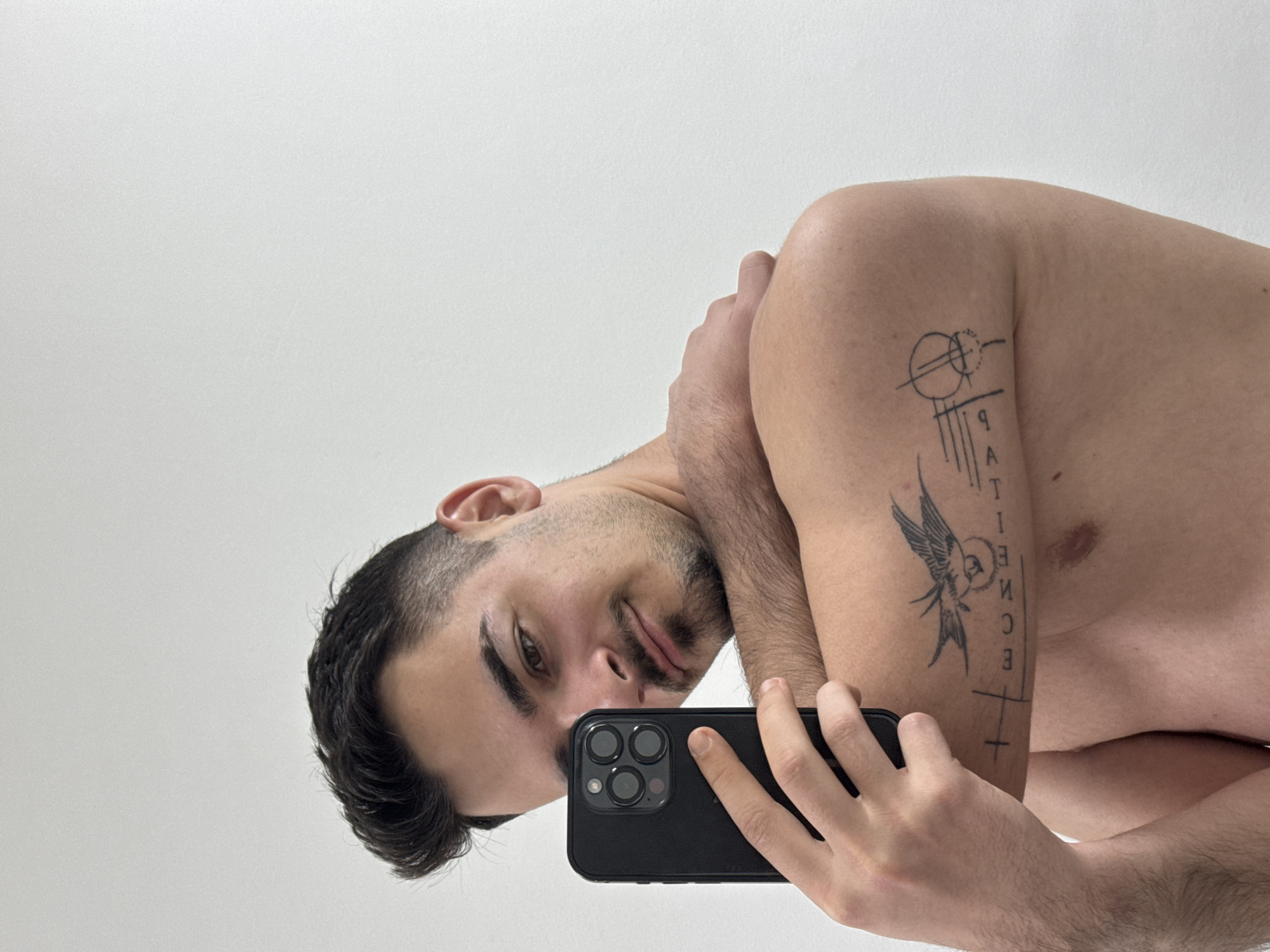 A light-skinned male is holding a black iPhone camera, facing downward, to take a selfie. He has dark hair, a trimmed beard and mustache, and visible chest hair. His right arm is bent and placed across his chest, revealing a tattoo on his upper arm consisting of abstract shapes, text, and a bird. The background is a plain, light wall. There are no indications of a location in Sofia, Bulgaria.