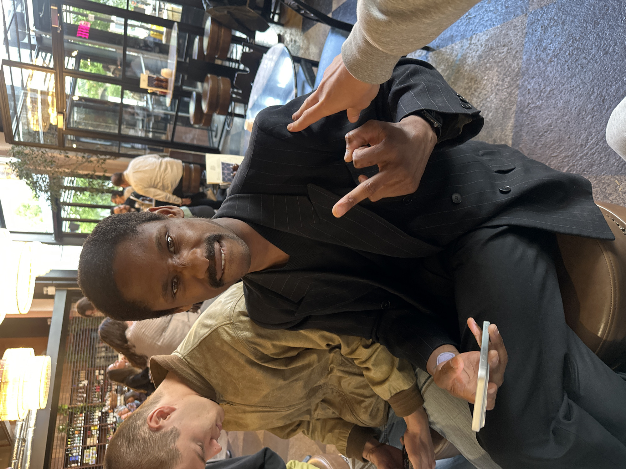 A vertical medium shot captures a man of color in a dark pinstripe suit jacket, seated in what appears to be an indoor restaurant setting. The man is smiling at the camera. Another hand with lighter skin is pointing toward the jacket. The man has a phone in his hand. In the background, one can see other patrons seated at tables. The setting appears to be a restaurant or bar in London, United Kingdom, identifiable by the distinct architectural style of the building and interior design.